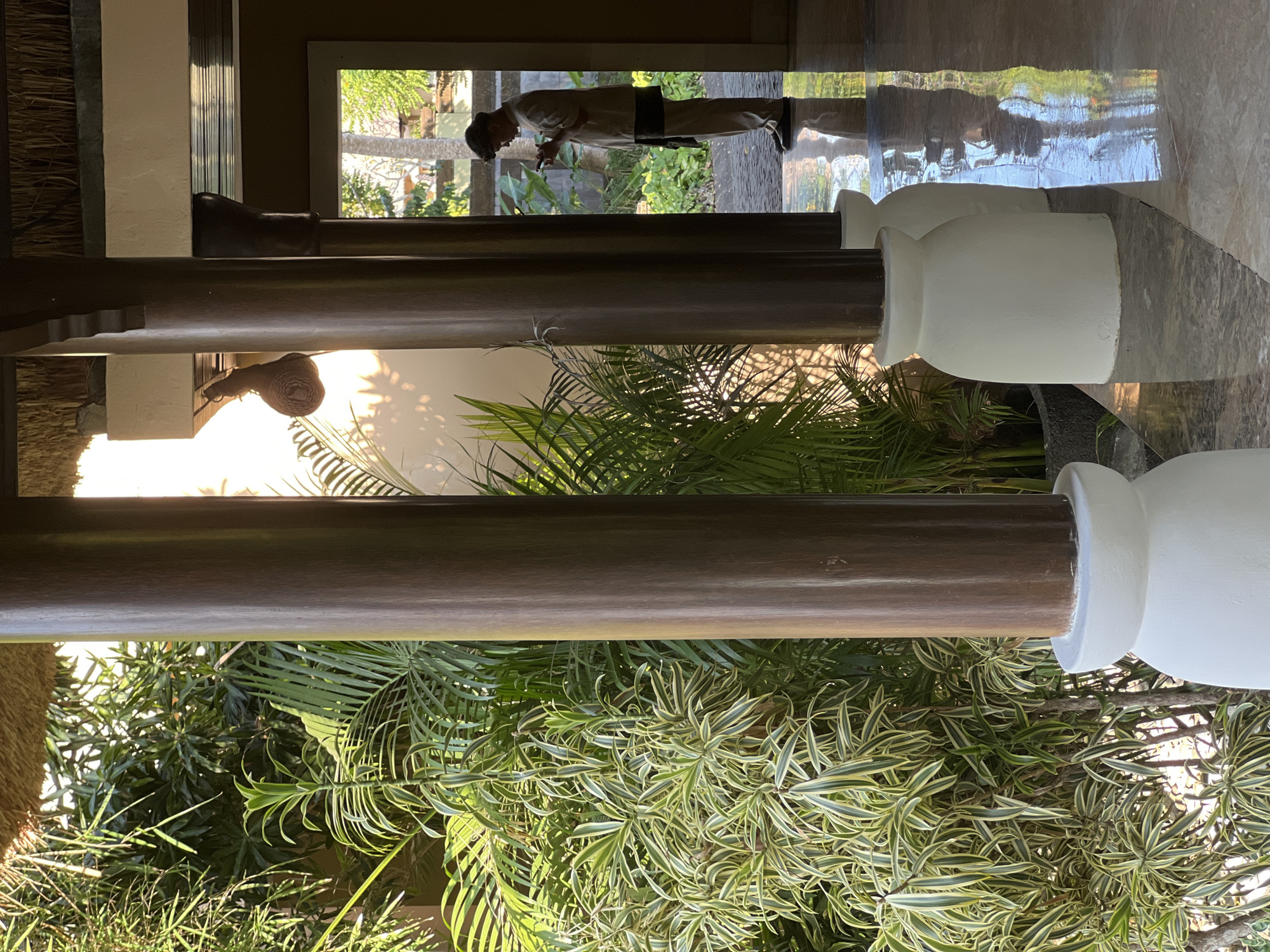 The image captures a view through an open-air walkway at a resort in Kuta Selatan, Indonesia. Two large, dark wooden pillars with white bases frame the scene. To the left, lush green foliage with variegated leaves is visible, catching the sunlight. To the right, a person dressed in a light-colored uniform and a cap stands in the opening of the walkway, looking down at a mobile phone. This individual is wearing a darker sash or sarong around their waist. The floor of the walkway is polished stone, reflecting the overhead structure, greenery, and sunlight. In the distance, through the opening, another palm tree and more greenery are visible. A woven decorative item hangs from the ceiling on the right side of the walkway. The overall impression is of a tropical setting with a blend of natural elements and architectural features.
