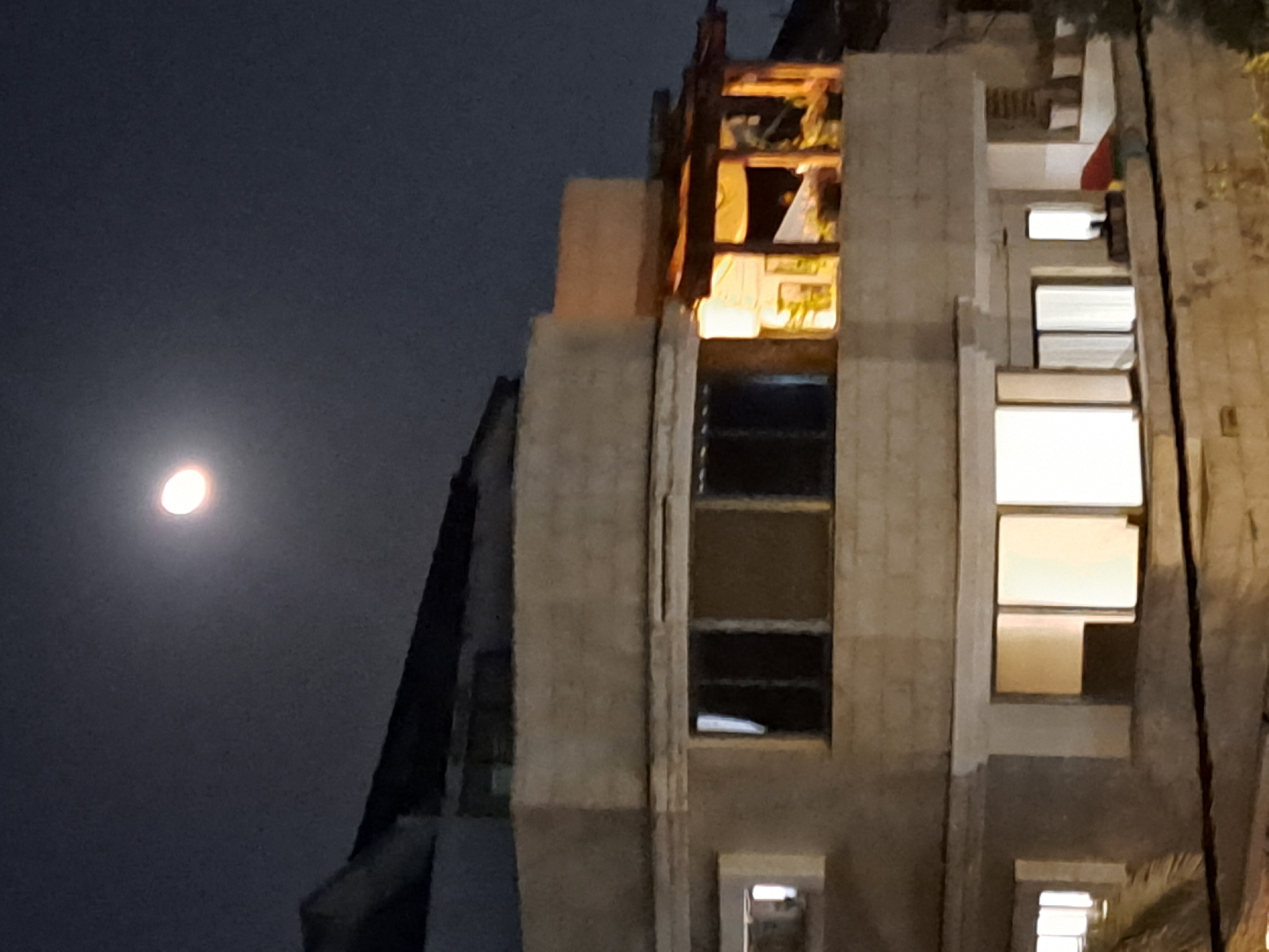 The image captures a nighttime scene featuring a partially eclipsed moon in a dark sky above a multi-story building. The building appears to be constructed from light-colored brick and concrete, with several windows visible. Some windows are illuminated from within, suggesting occupancy or lighting within the structure. There is a covered balcony area on an upper floor with lights on, revealing plants and furniture. The lower portion of the image shows more windows, some with internal lighting. The overall impression is of an urban or suburban residential setting in Ramat HaSharon, Israel, under a night sky.