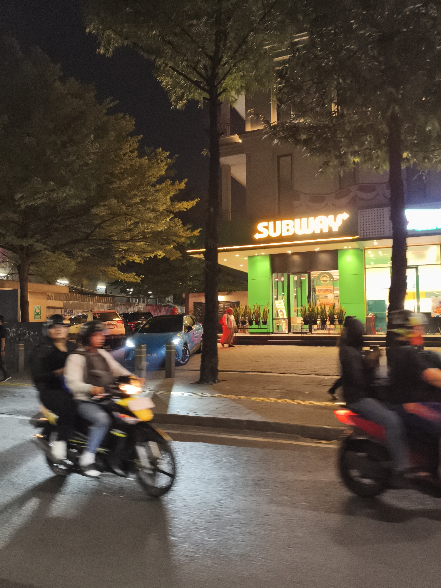 The image depicts a street scene at night in South Jakarta, Indonesia, with the prominent presence of a brightly lit Subway restaurant. Two individuals are riding on a yellow motorcycle in the foreground, moving from left to right and appearing blurred due to motion. The rider wears a helmet and a grey jacket, while the passenger behind them is dressed in black. A second motorcycle, predominantly red, is visible further to the right, also in motion, with two blurred figures riding it. Several cars are parked or driving on the street behind the motorcycles. A blue car with a distinctive graphic design is noticeable. The Subway restaurant features its signature green facade and illuminated orange "SUBWAY" signage. Trees with full foliage line the street, and the overall lighting suggests an urban environment at night. The ground is paved, and a sidewalk is present.