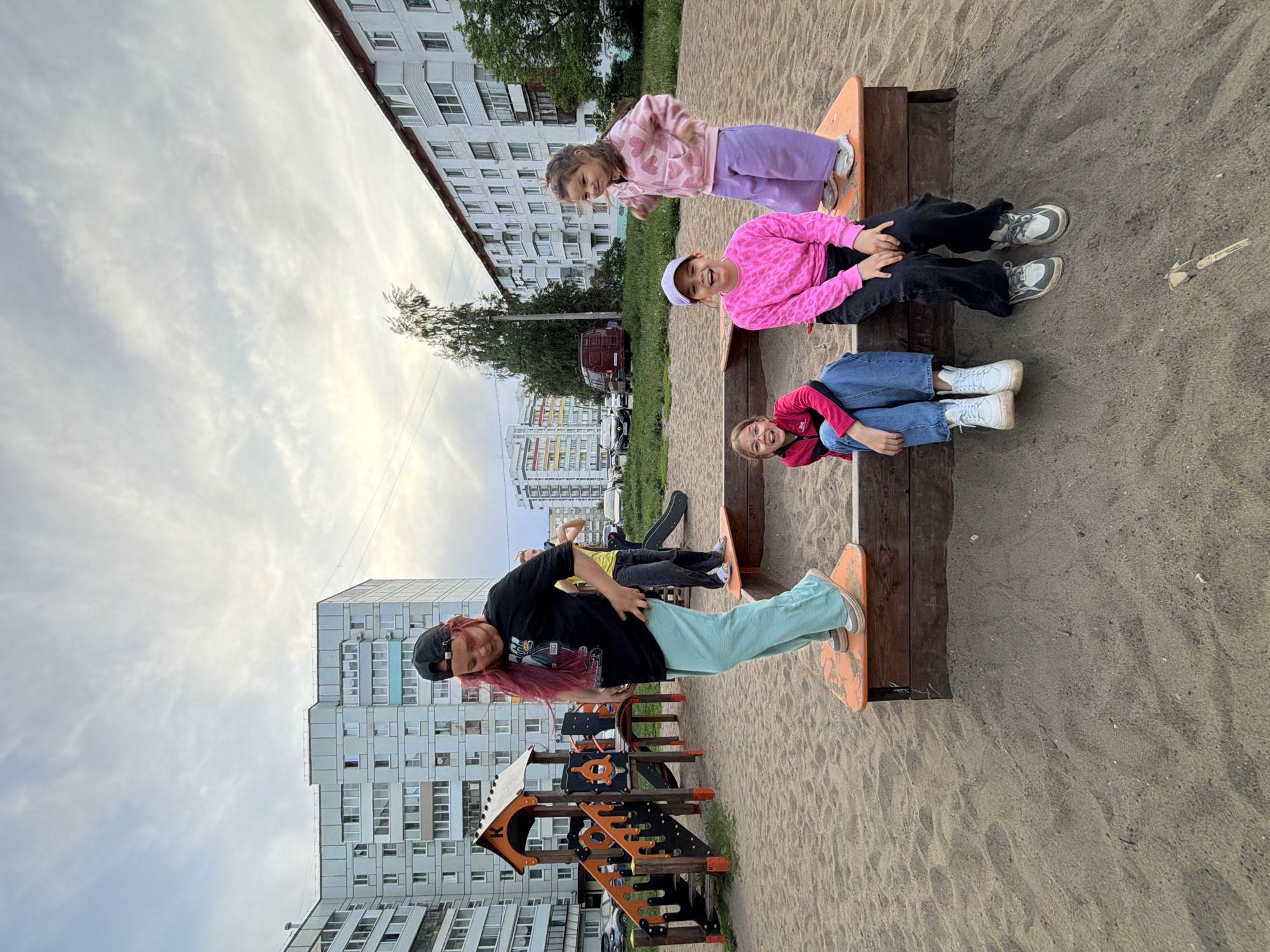 Four girls are positioned on a seesaw in a sandy playground. The first girl has pink hair, wears a black shirt and teal pants, and stands on one end of the seesaw. The second girl wears a yellow shirt, black pants, and sneakers. The third girl wears a pink shirt with a white hat, black pants and sneakers. The fourth girl wears a red shirt and blue jeans and white sneakers. The playground is located in the city of Chaikovsky, Russia with apartment buildings visible in the background. The sky is mostly cloudy.