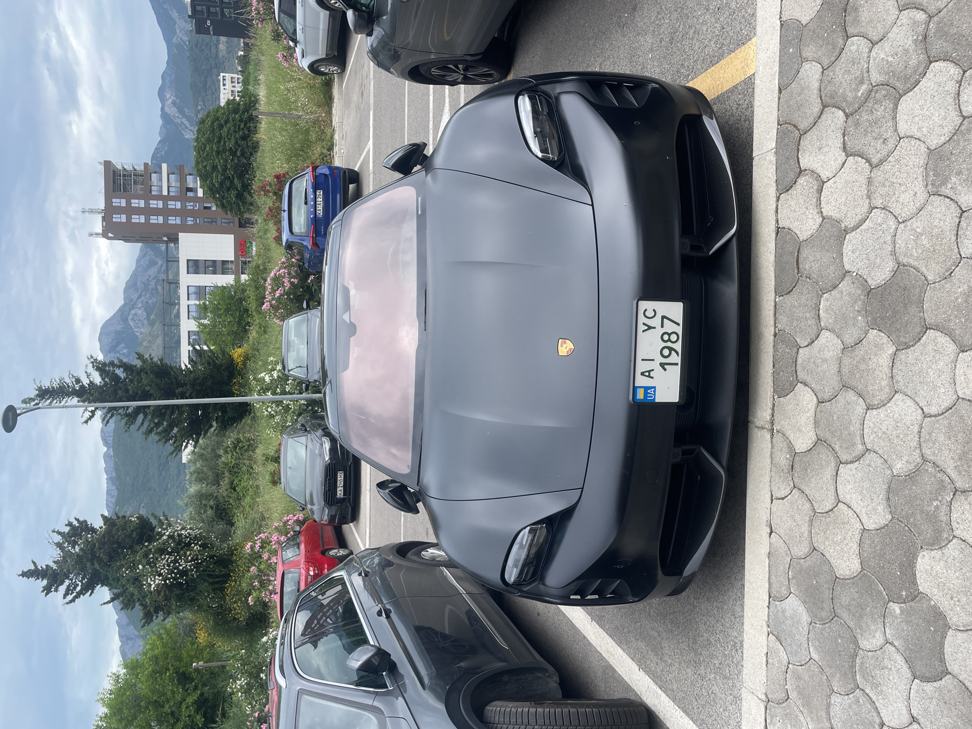 A dark-colored Porsche Taycan, parked head-on between two other cars in a parking lot, is the focal point. The car's front license plate reads "AI YC 1987" with a Ukrainian flag. The parking lot is paved, with white lines marking parking spaces and a yellow line separating spaces from a sidewalk with gray cobblestone. A landscaped area behind the parking spaces contains trees, shrubs with pink flowers, and a light pole. A multi-story building and mountains are visible in the background. The location is in the city of Bar, Montenegro.