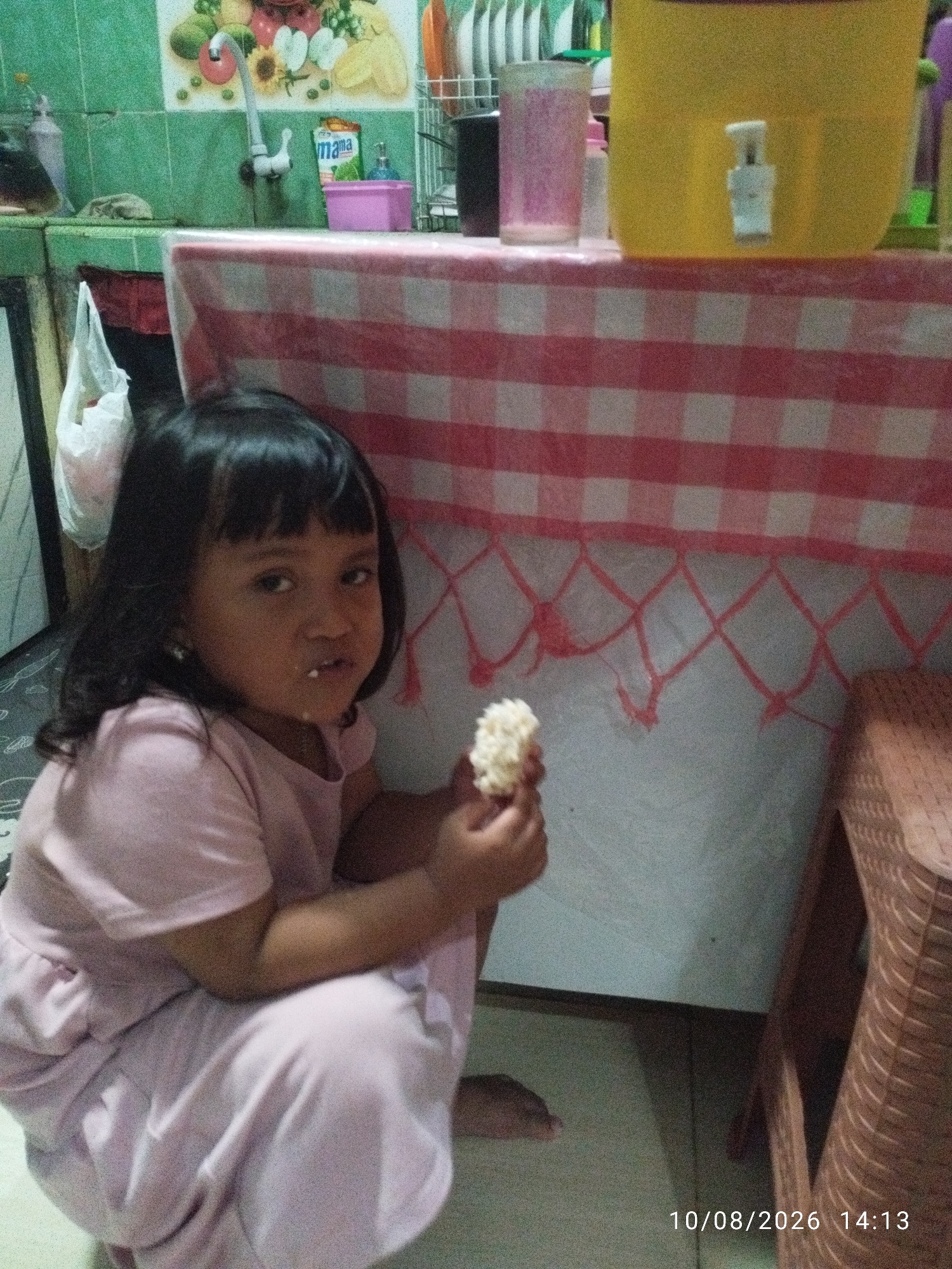 An outdoor shot of a young girl, likely Indonesian, squatting and holding a small piece of food in her right hand. The girl is wearing a pink shirt and pants. She has dark hair and dark eyes. The background features a kitchen counter with a red and white checkered tablecloth, a yellow water dispenser, and a tile backsplash. The timestamp at the bottom of the image indicates the date is October 8, 2026, and the time is 2:13 PM. The image appears to have been taken in an urban or suburban setting within Bandar Lampung, Indonesia, based on the typical kitchen design and the presence of the Indonesian language on a product in the background.