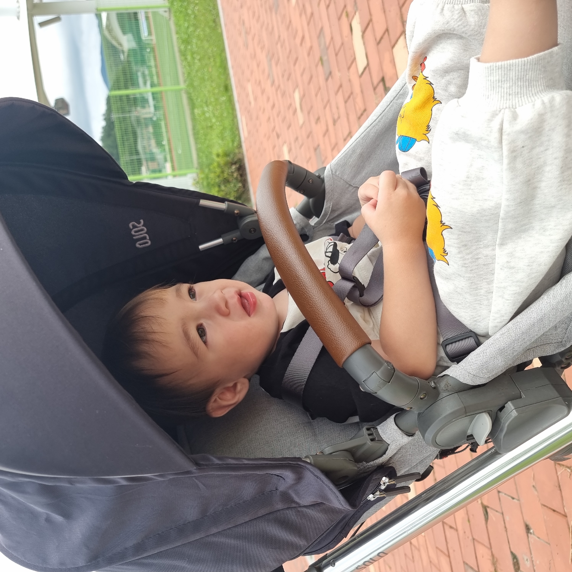 A young child, likely under three years old, is seated in a stroller outdoors. The child has light brown hair, dark eyes, and is looking to the right with their mouth slightly open. They are wearing a black and white shirt with a graphic print and light gray pants with a yellow cartoon character design. The stroller is dark gray with a brown handlebar. The background shows a paved walkway made of red bricks, a green fence, a grassy area, and buildings in the distance. The sky is overcast. The location is identified as Dunnae-myeon, South Korea.
