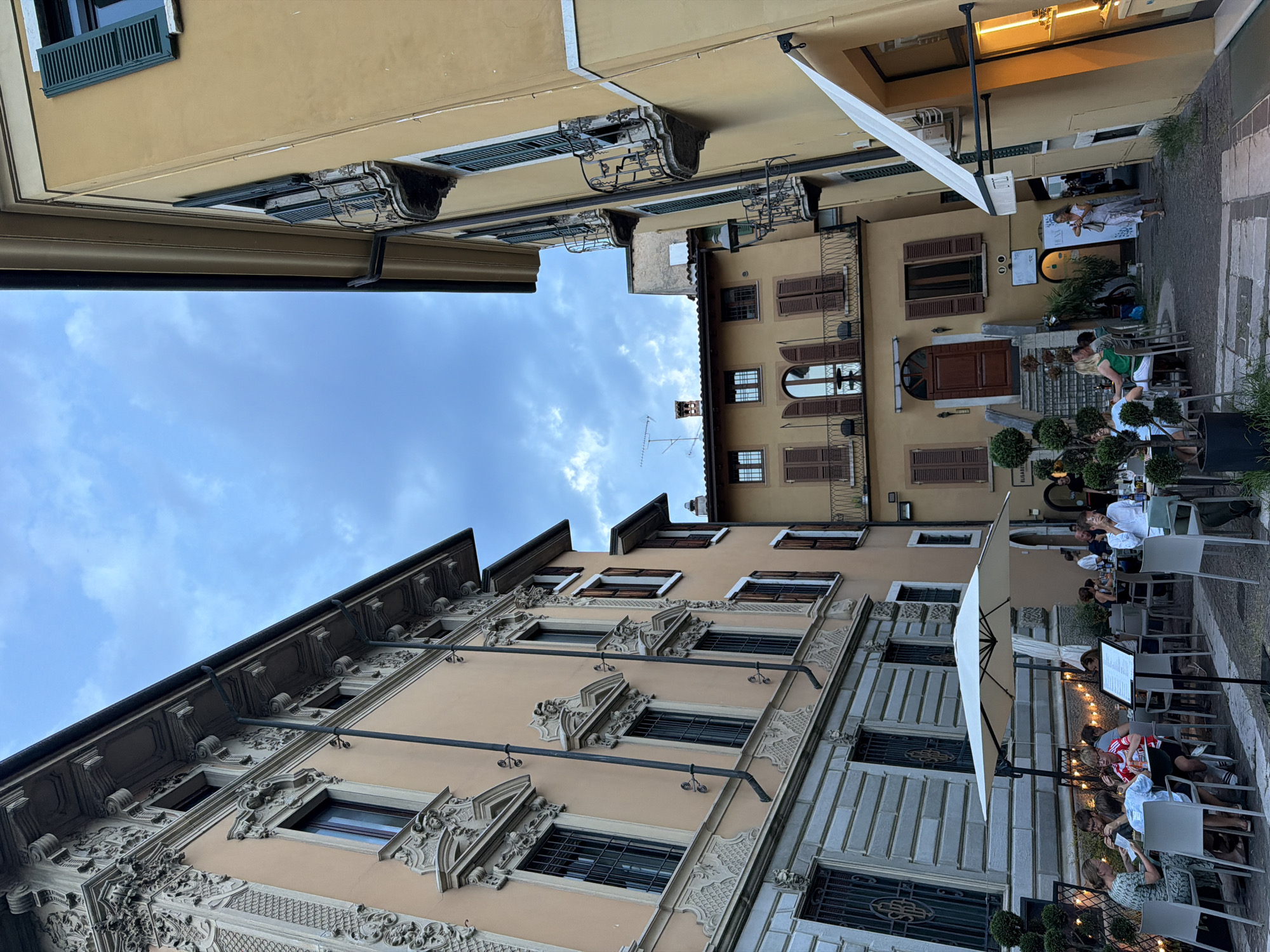 An outdoor dining area is set up in front of ornate buildings in Salò, Italy. In the foreground, a large white umbrella shades a group of people dining at a table. A menu board stands beside them. Several other tables with patrons are visible in the background, as are the exteriors of buildings with architectural details, balconies, and shutters. The scene is lit by string lights and a warm interior light from a building on the right. The sky is overcast with clouds.