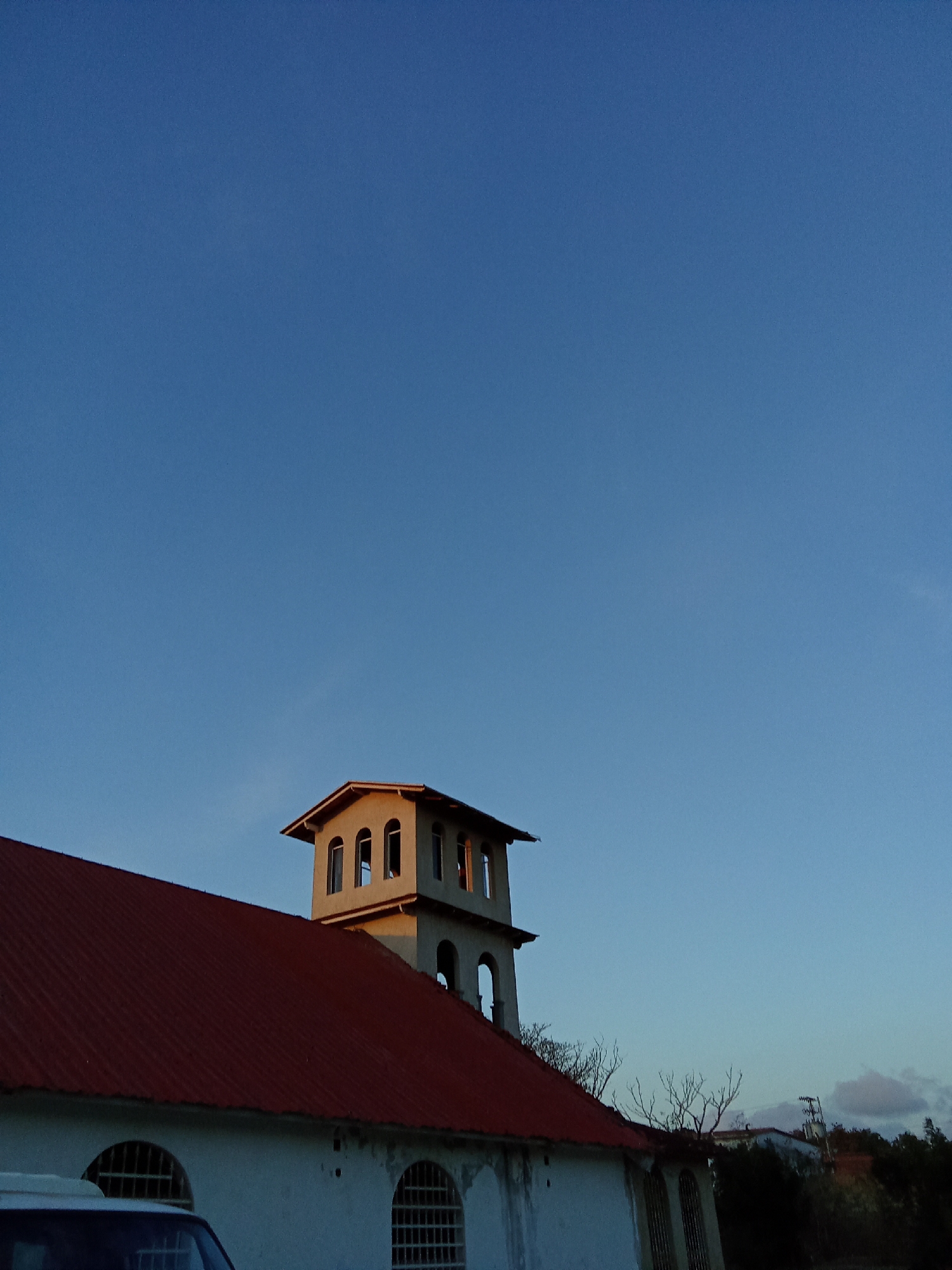 The image captures a building in Juan Griego, Venezuela, under a clear blue sky transitioning towards dusk. The building is predominantly white with a red roof and a distinctive tower structure. The tower has multiple arched windows. The architecture exhibits signs of age and possible disrepair. A portion of a vehicle is visible in the foreground. In the background, sparse trees and a glimpse of other buildings and utility poles can be seen on a distant hill. The lighting suggests either early morning or late afternoon. No people or interactions are visible in the scene.