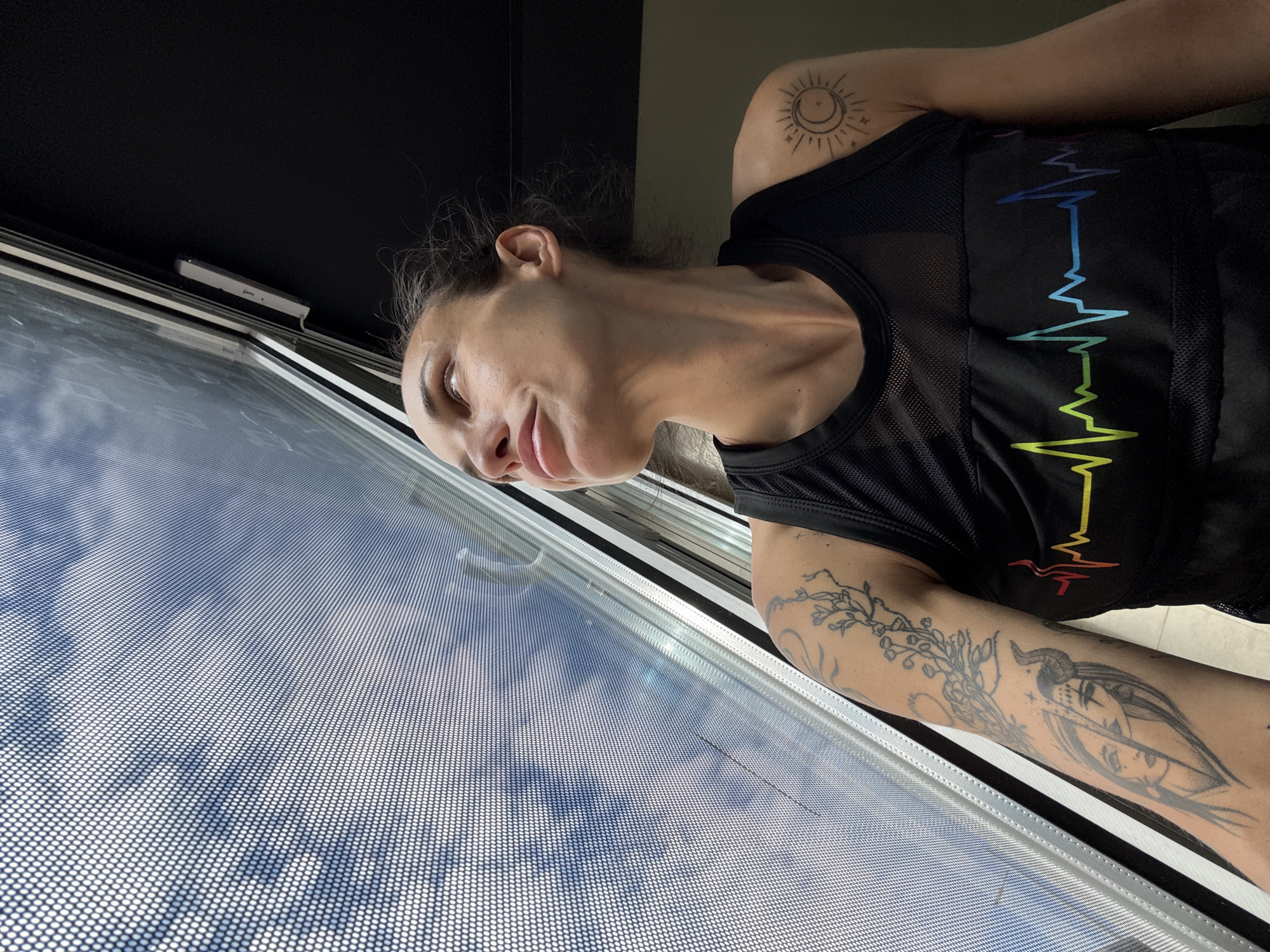 A person with tattoos stands near a window. The person is wearing a black tank top with a rainbow-colored electrocardiogram-style design across the chest. The person has a tattoo of a sun/moon on their upper right arm and a tattoo with a woman with a horned headress and branches on their lower right arm. The window has a screen and appears to be showing a view of the sky and clouds. The background is black. No location information about Pleven, Bulgaria is discernible from the image.
