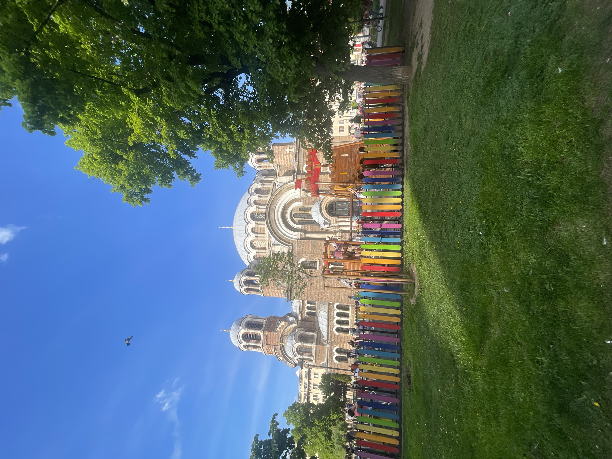 A park scene in Sofia, Bulgaria, shows the Alexander Nevsky Cathedral behind a colorful fence. The fence is composed of vertical planks painted in various colors, arranged in a line along the edge of the park. The green grass extends from the fence towards the right side. To the left, the view is dominated by the cathedral, with its domes and arches visible against the blue sky. A green tree overhangs the top-left portion of the scene. A few people are visible on the playground behind the colorful fence. A flying bird is seen in the blue sky.