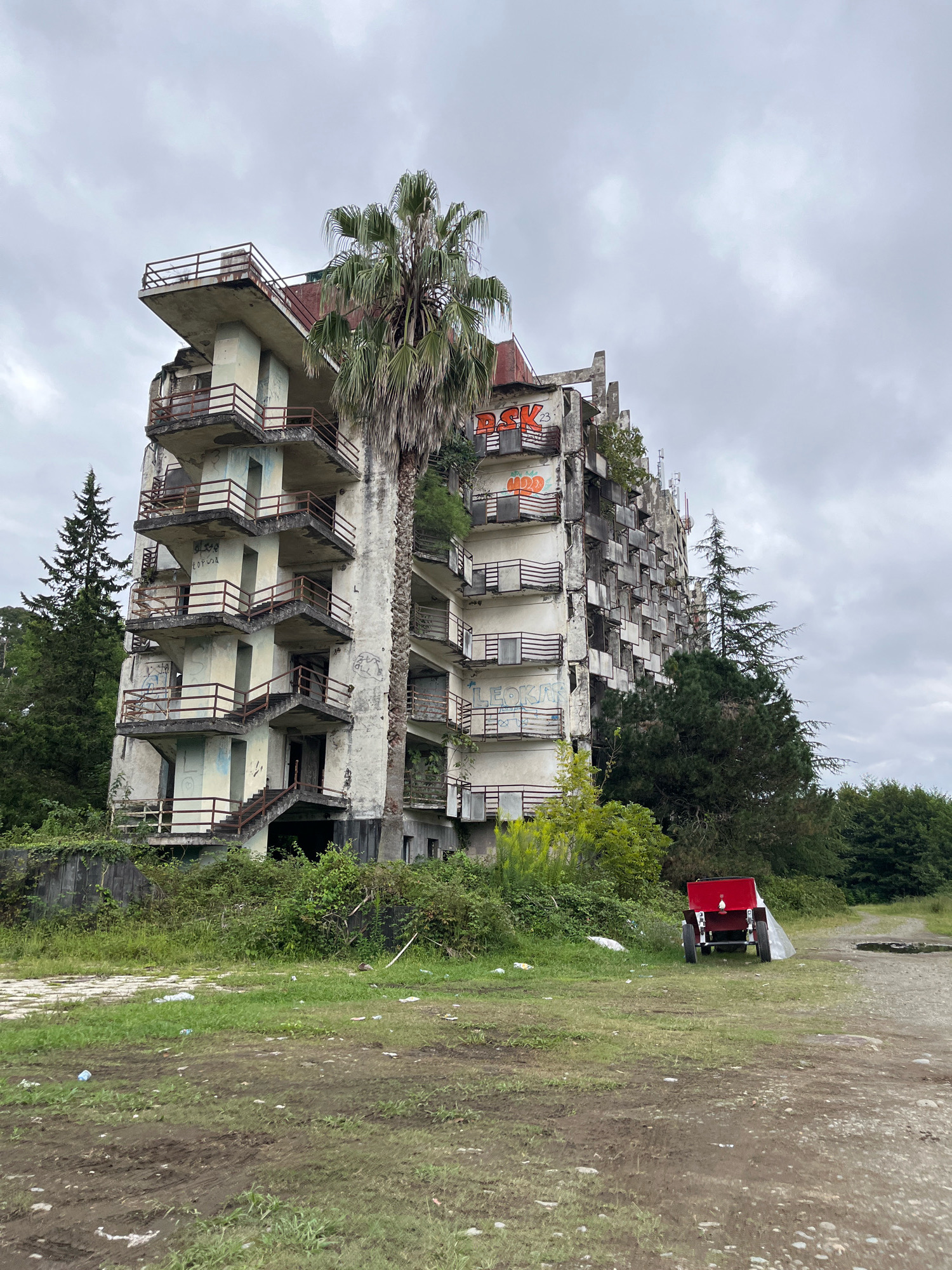 The image shows an abandoned, multi-story building in Ureki, Georgia. The building appears to be a former hotel or apartment complex, with balconies and open doorways visible. It is in a state of disrepair, with peeling paint, graffiti, and overgrown vegetation. A tall palm tree stands next to the building, partially obscuring its facade. In the foreground, there is a red trailer with two wheels, parked on a dirt and grass area. The sky is overcast, suggesting a cloudy day. The surrounding environment consists of trees and foliage, indicative of a natural setting encroaching on the derelict structure.