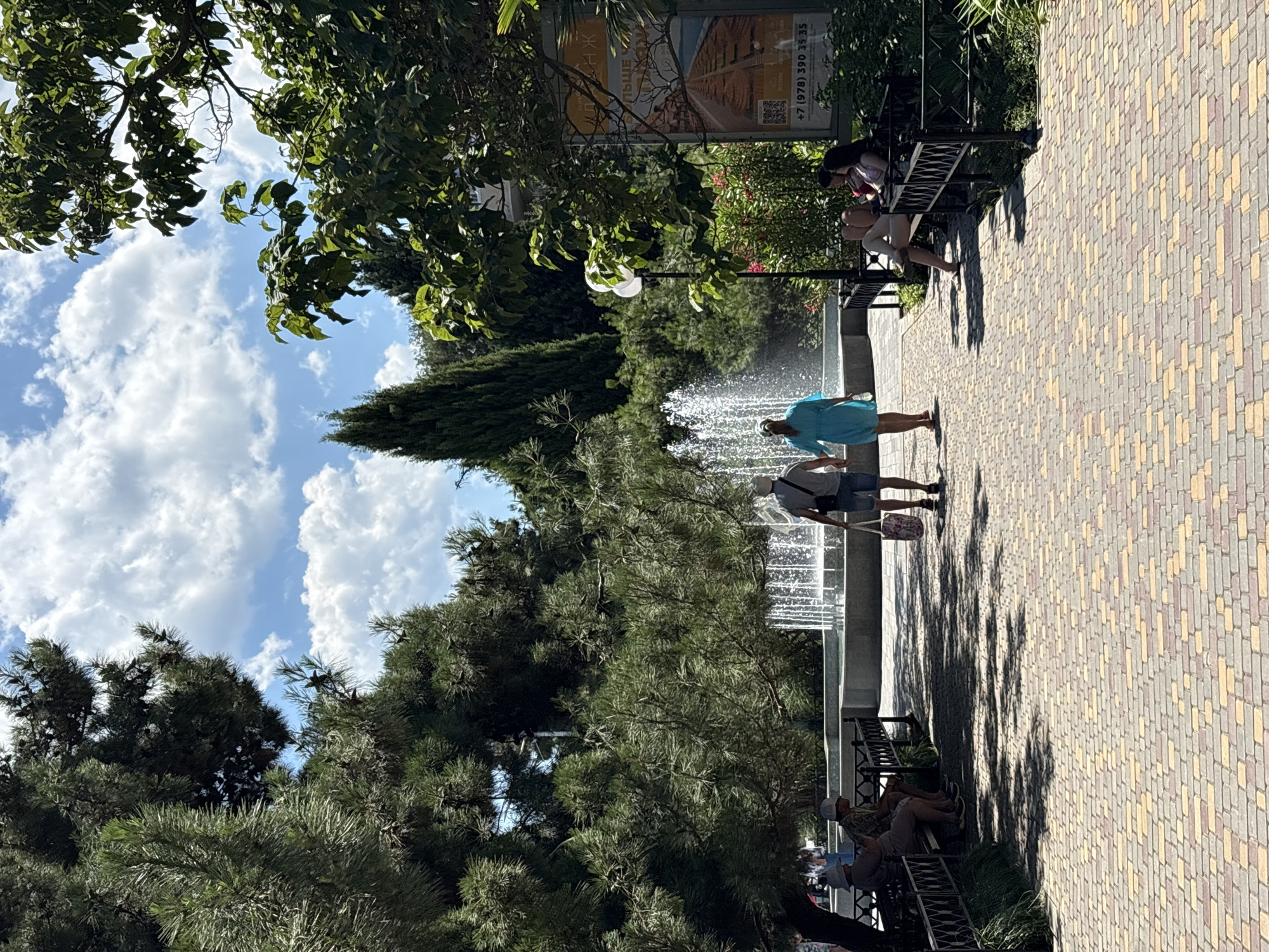 In Alushta, Ukraine, a scenic park features a paved walkway lined with benches and lush greenery. In the foreground, two individuals stroll hand-in-hand along the walkway, with one carrying a patterned bag. To their left, two people sit on a bench, and further left, more individuals are visible on another bench. The background is dominated by a large fountain spraying water, surrounded by trees and a tall, conical evergreen tree. The sky above is a bright blue with scattered white clouds. A poster is affixed to a lamppost on the right side of the image.