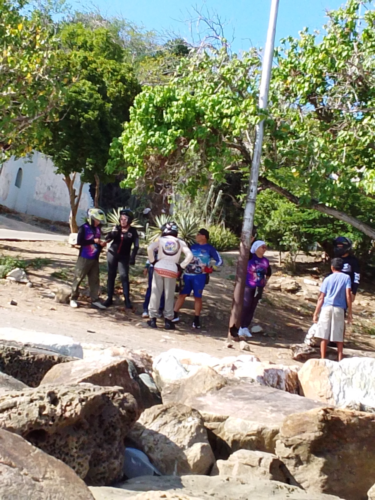 The image depicts a group of individuals gathered on a dirt path near a rocky shoreline in Pedro González, Venezuela. The environment is characterized by lush green trees, a bright blue sky, and several large rocks in the foreground.
There are at least eight people visible in the scene. Several individuals are wearing helmets and what appear to be protective gear, suggesting they might be involved in an activity like motorcycling or cycling.
One person on the left is wearing a colorful long-sleeved shirt and green pants, with a yellow helmet. To their right, a person in black with a black helmet and dark pants is standing. Further to the right, a person in a white top with colorful accents and grey pants, wearing a helmet, is facing away from the camera. Next to them, a person in a blue shirt and white shorts is facing the camera. Another individual in a purple shirt and dark pants is standing near a wooden pole. To the far right, a person in a black shirt with "FIX" written on it is facing a young person in a blue shirt and white shorts. Another person in a blue shirt and blue shorts is partially visible in the background.
The overall scene appears to be a casual gathering in an outdoor, possibly recreational, setting. The presence of protective gear implies an activity is either ongoing or about to commence.