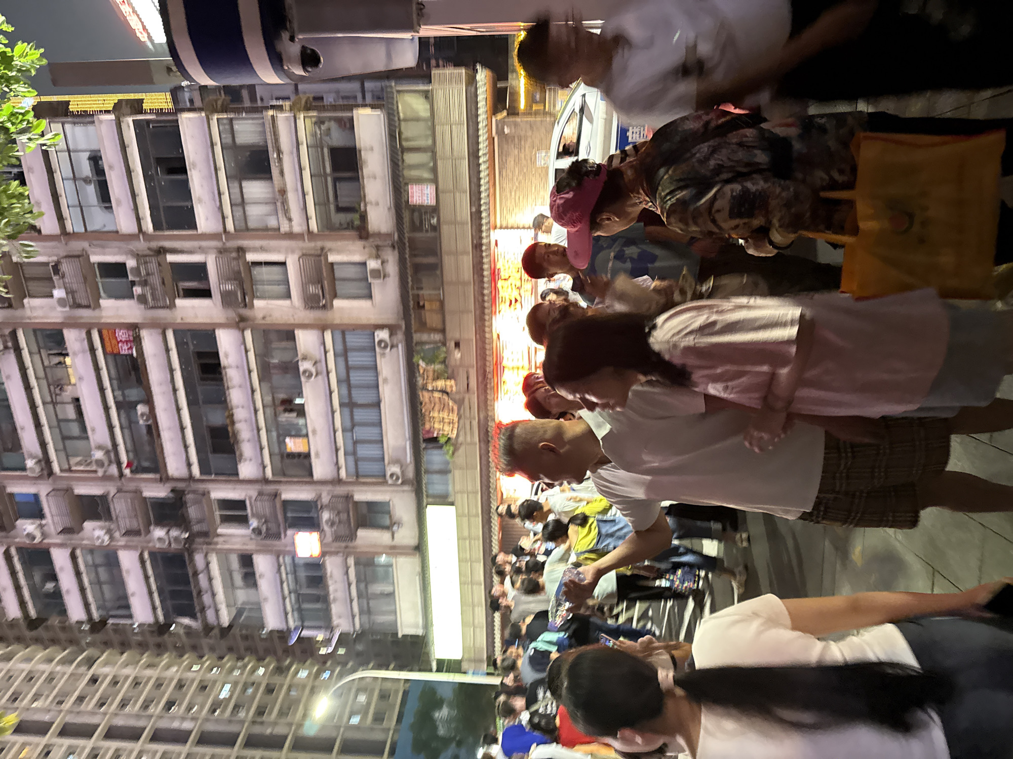 A crowded street scene at night in Yuzhong District, China features a diverse group of people gathered in front of a multi-story building. The building, with its numerous windows and balconies, is illuminated by artificial lights. Some windows display signs, including one with red lettering. In the foreground, several individuals are visible, including a man in a white t-shirt and plaid shorts holding a clear plastic bottle, and a woman in a pink shirt. A yellow shopping bag is prominent in the lower right portion of the image, held by a person in a patterned jacket. The street is paved, and streetlights cast a glow on the scene. A white car is partially visible in the background, and a security camera is mounted on a pillar. The overall atmosphere suggests a lively evening gathering or public space.