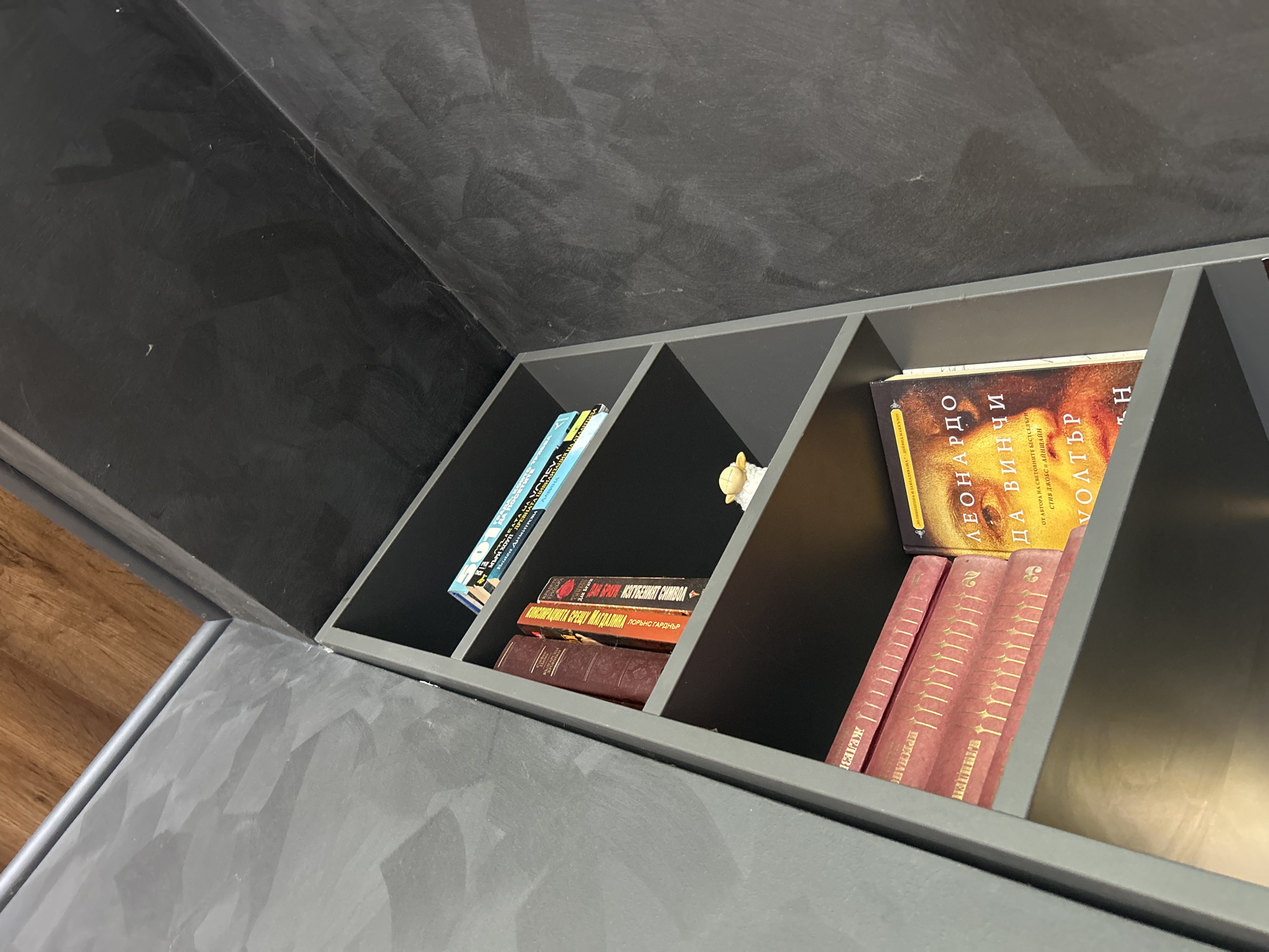 An interior scene in Unknown, Unknown, captured from a low angle, depicts a dark grey, multi-shelf bookcase integrated into a corner of a room. The walls are finished in a dark grey plaster or paint with visible lighter brushstrokes, creating a textured, mottled appearance. A wooden floor with a grey metallic trim is visible at the top of the frame, indicating an upper level or a boundary with a different surface.
The bookcase itself is dark grey and contains various books. On the top shelf, several horizontally stacked books are visible, including one with a blue and white cover featuring Cyrillic text, partially readable as "101 СЪВЕТА НА НА ТААЛИАНА АНТОНИ ДИМИТРОВ". The middle shelf holds several vertically oriented books; a dark red book with gold spine lettering is partially obscured, followed by a red book titled "ИСТОРИЯТА СРЕЩУ МАДАЛИНА ПОРЧА И АРТУР" in Cyrillic, and another red book. A small, light-colored figurine, possibly a sheep, stands between two books on this shelf. The bottom shelf, partially visible, displays several red books with gold decorative spines, horizontally stacked, with "КЕЙДЖ" visible on one spine. Next to them, a vertically placed book with an orange and yellow cover clearly displays the Cyrillic title "ЛЕОНАРДО ДА ВИНЧИ" and "ВОЛТЕР" below it, with additional text indicating it is "ОТ АВТОРА НА СВЕТОВНИТЕ БЕСТСЕЛЪРИ СТИВ ДЖОБС И АЙЗЪКСОН". The overall environment is characterized by a muted color palette dominated by greys and dark tones, contrasted by the colors of the books and the warm tone of the wooden floor. No individuals are present in the image.