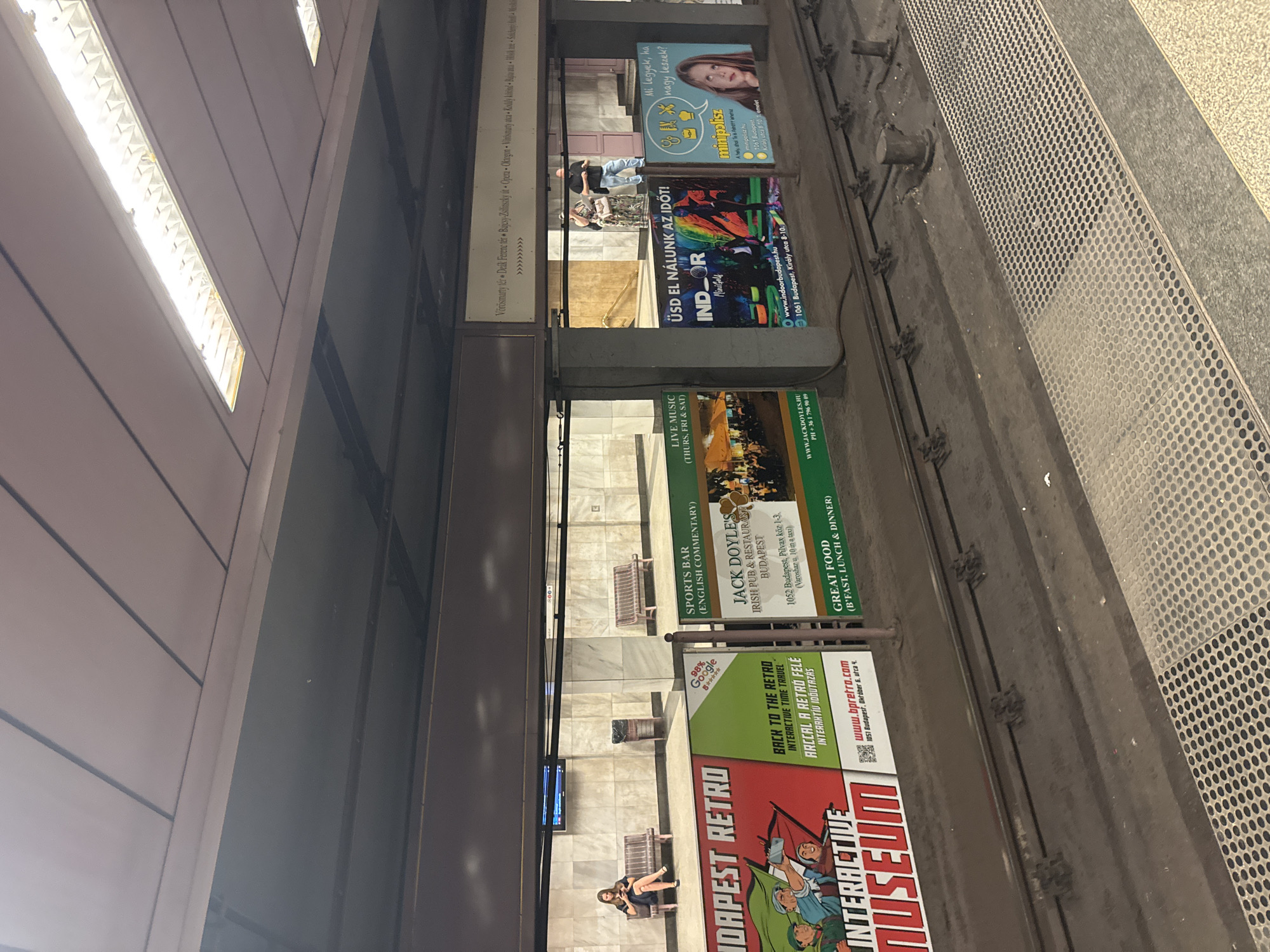 The image shows a view of a Budapest metro station platform. In the foreground, there are train tracks and a textured metal grating along the edge of the platform. Various advertisements are displayed on panels along the station wall.
On the left, a large advertisement for "BUDAPEST RETRO INTERACTIVE MUSEUM" is visible, with colorful illustrations. To its right, smaller ads include "BACK TO THE RETRO INTERACTIVE TIME TRAVEL" and information for "JACK DOYLE'S IRISH PUB & RESTAURANT," which also advertises "GREAT FOOD." Further to the right, there's an ad for "INDOOR MiniGolf" and another advertisement featuring a young girl and Hungarian text.
In the background, a woman sits on a bench, looking at her phone. Another bench is visible further down the platform, along with a trash can. Several pillars support the ceiling structure. The walls are tiled with a marble-like pattern. The ceiling features fluorescent lighting panels. A directional sign with Hungarian place names is mounted above the advertisements. Two people are standing near a staircase in the background, to the right of the center.