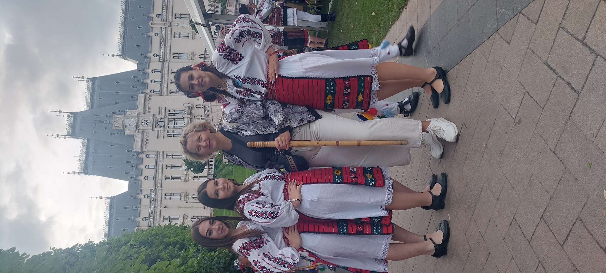 This image captures a group of four individuals, three young women and one adult woman, posing outdoors. The three young women are dressed in traditional Romanian folk costumes, characterized by white blouses with intricate red and black embroidery on the sleeves and bodices, and red skirts adorned with similar patterns. They are wearing dark, flat shoes. The adult woman, positioned between the young women, is wearing a grey and black patterned jacket, white pants, and white sneakers. She is holding a light-colored wooden staff. They are standing on a paved area in front of the Palace of Culture in Iași, Romania. The Palace, a grand architectural landmark with multiple spires and ornate detailing, is visible in the background under a cloudy sky. Green trees are present on the left side of the frame, and a white tent structure can be seen to the right. The overall scene suggests a cultural event or performance taking place in a public space.
