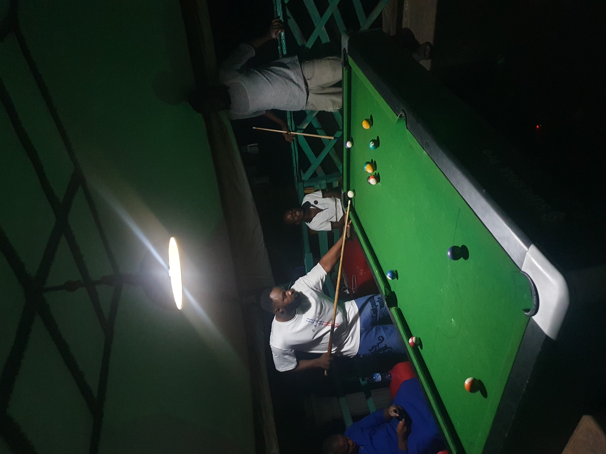 In the city of Lokoja, Nigeria, four individuals are gathered around a green billiard table. The scene is dimly lit, with a single overhead light illuminating the table and its surroundings. Three men and one woman appear to be playing or observing a game of pool. One man, wearing a white t-shirt and blue shorts, is actively taking a shot with a cue stick. Another man, in a checkered shirt and light-colored shorts, stands behind him, also holding a cue stick. A third person, a woman, is seated to the left of the table, dressed in a blue outfit, and appears to be using a mobile device. A young boy in a white polo shirt is standing at the far end of the table, holding a cue stick and looking towards the game. Several billiard balls are scattered across the green felt surface. The environment suggests an indoor recreational space, possibly a bar or club, with a ceiling structure and a green fence-like barrier visible in the background.