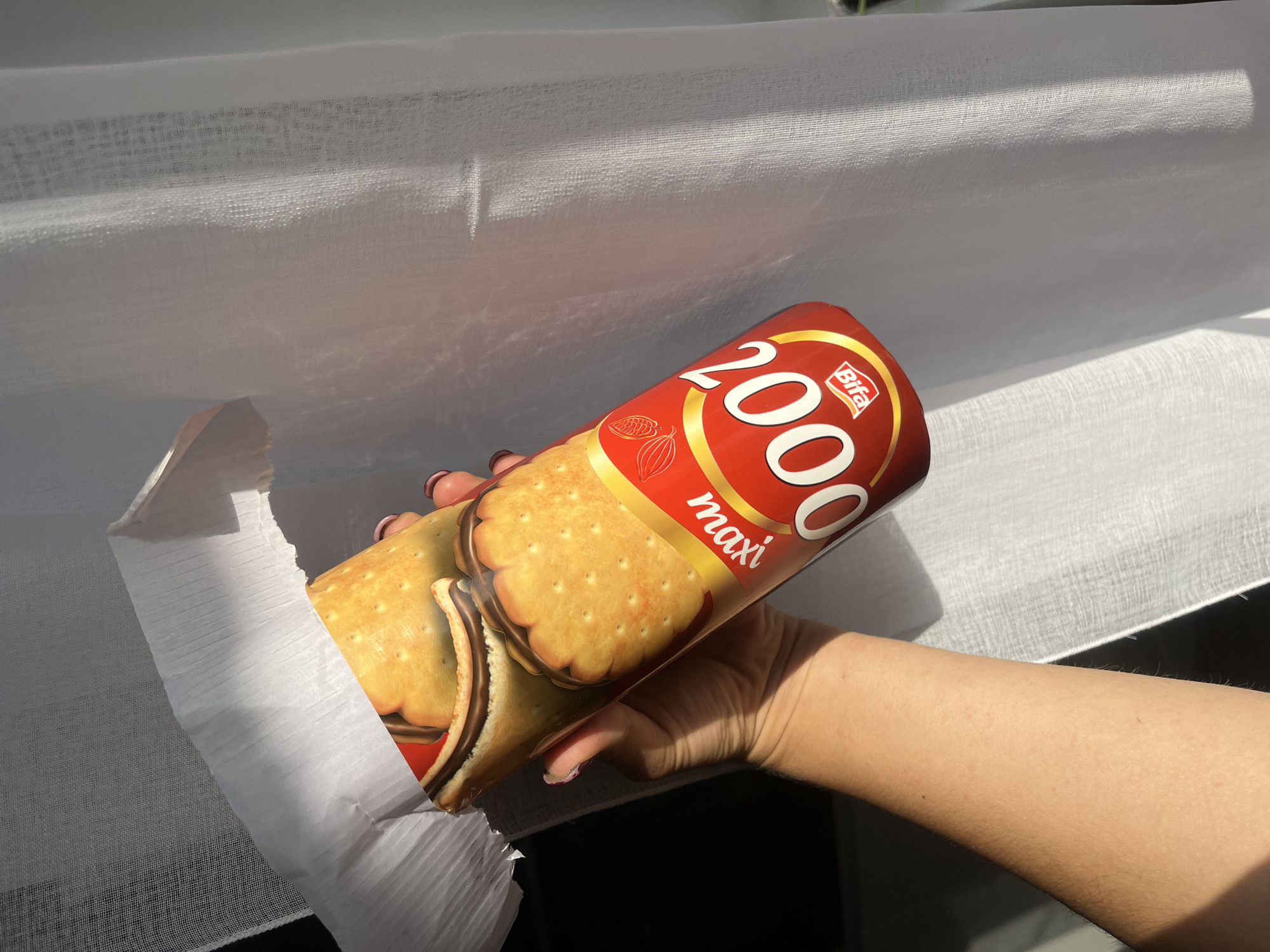 A person's arm and hand are visible in the foreground, holding a cylindrical package of "2000 Maxi" chocolate sandwich cookies. The package is red and yellow, featuring images of the cookies. The cookies themselves are golden brown with a ridged edge and a dark chocolate filling. The background consists of a sheer white curtain, illuminated by sunlight, casting shadows and creating a textured effect. The image is taken from a slightly tilted angle, with the package angled away from the viewer. There is no discernible urban or natural landscape visible, making it impossible to determine a specific location within Сеніцкі сельскі Савет, Belarus.