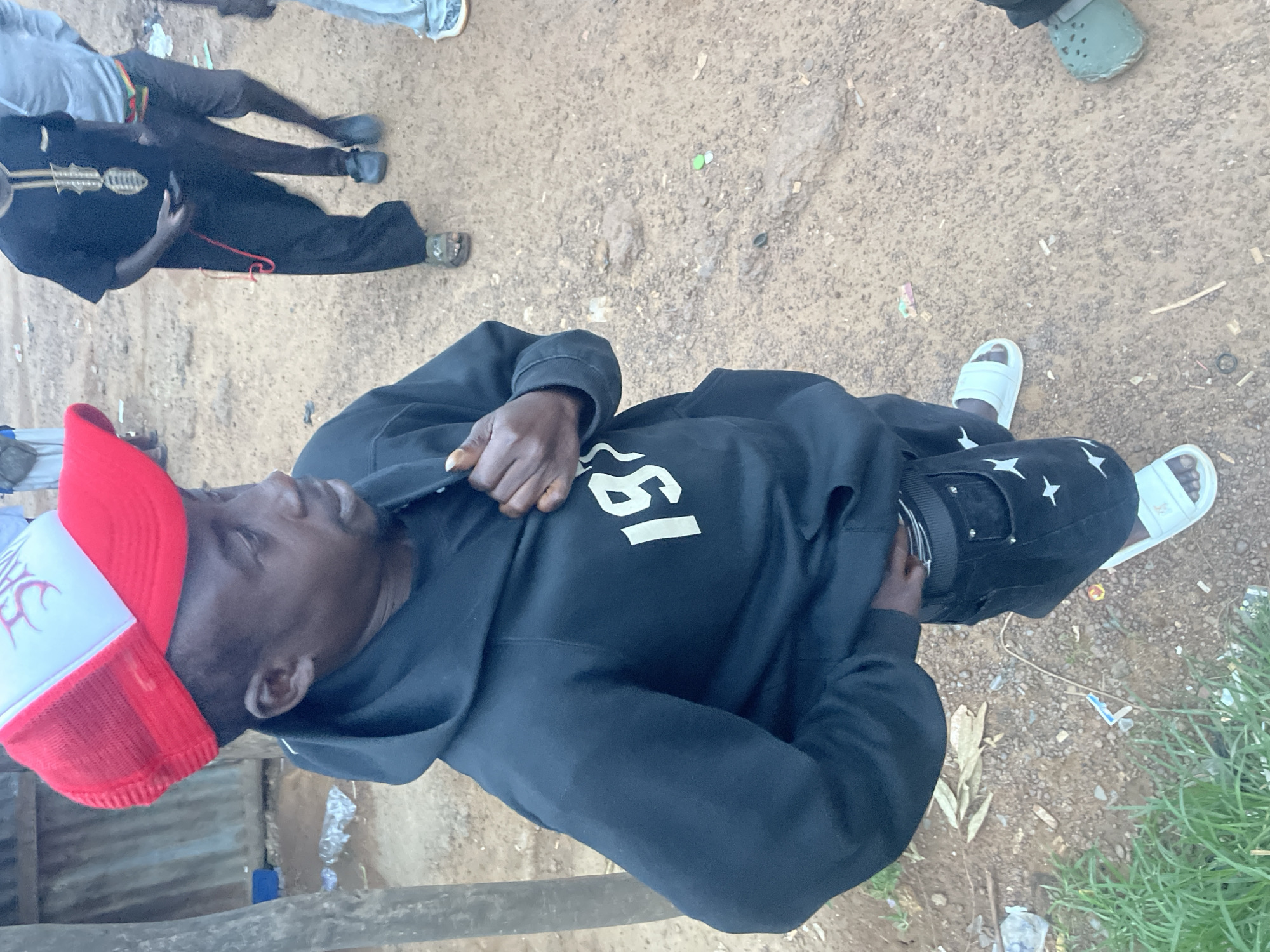 A man wearing a red and white baseball cap, a black hoodie with white lettering, black jeans with white star patterns, and white sandals stands on a dirt and gravel ground with some scattered debris and green grass. He appears to be pulling up his hoodie. In the background, other people are visible, some standing and some seated. The environment includes glimpses of what appear to be simple structures. The location is in Durimi, Nigeria.