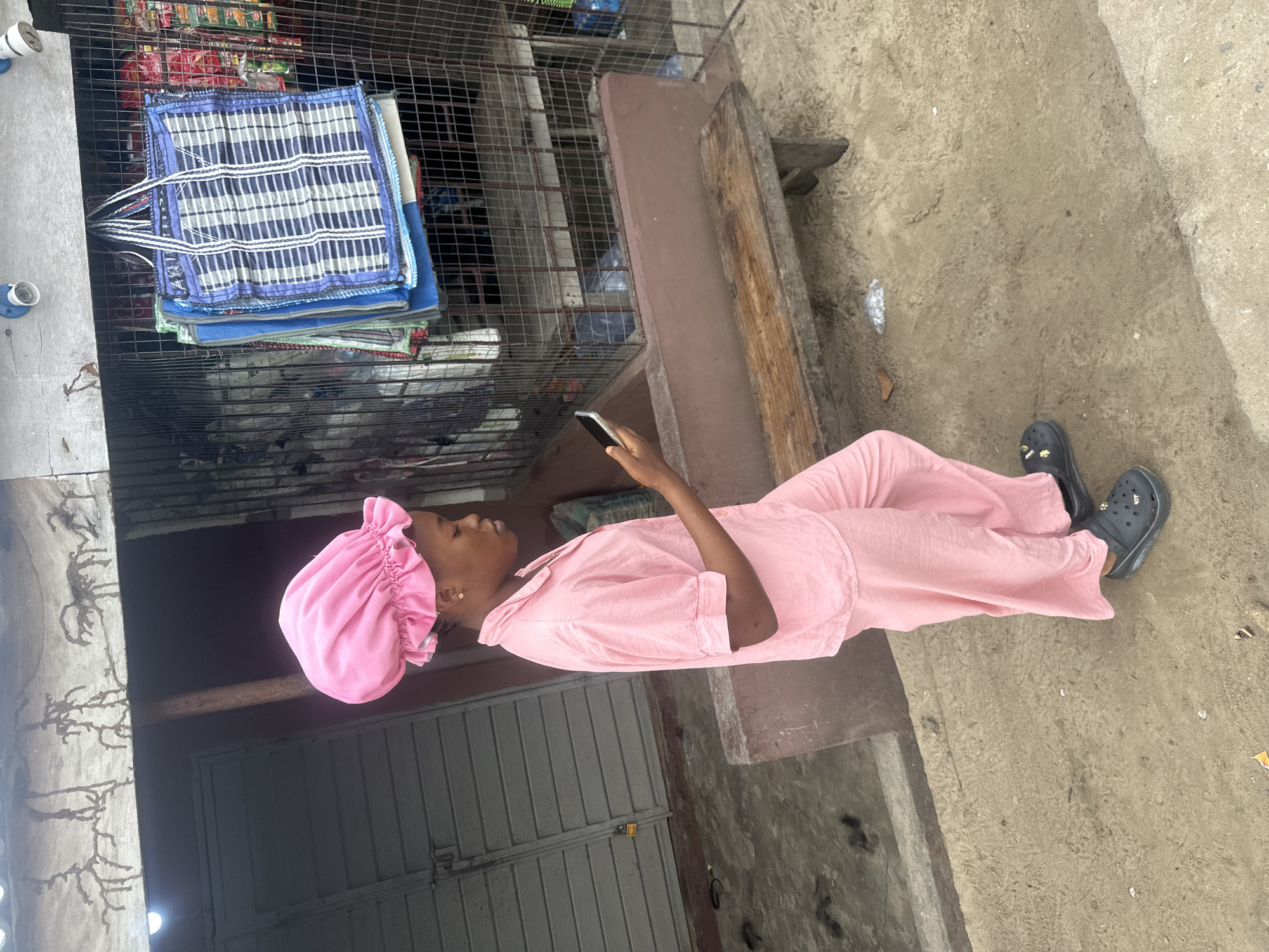 A young woman, wearing a pink bonnet and pink pajamas, stands outside a shop in Badagry, Nigeria, looking at her phone. The shop has a wire mesh front, displaying bags and various items for sale. A wooden bench sits in front of the shop. The ground is unpaved, with a concrete barrier to the right of the frame. The wall behind the woman has an unusual textured pattern.