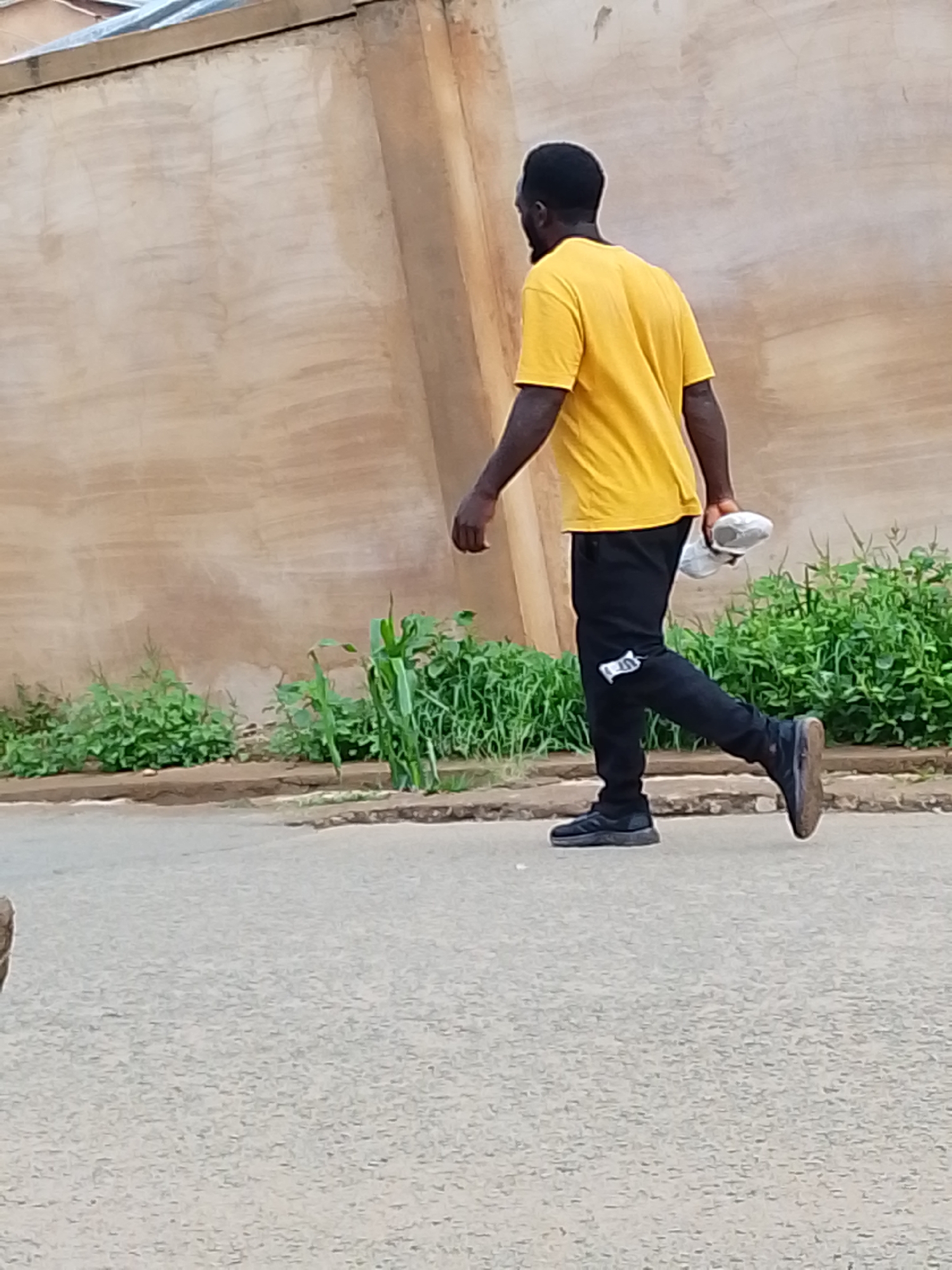 A man wearing a yellow t-shirt and black pants is walking on a paved road next to a sidewalk with green plants growing along a wall. He is carrying a pair of white sneakers in his hand. The location appears to be in a city based on the presence of the road and sidewalk, but the specific city is unknown.