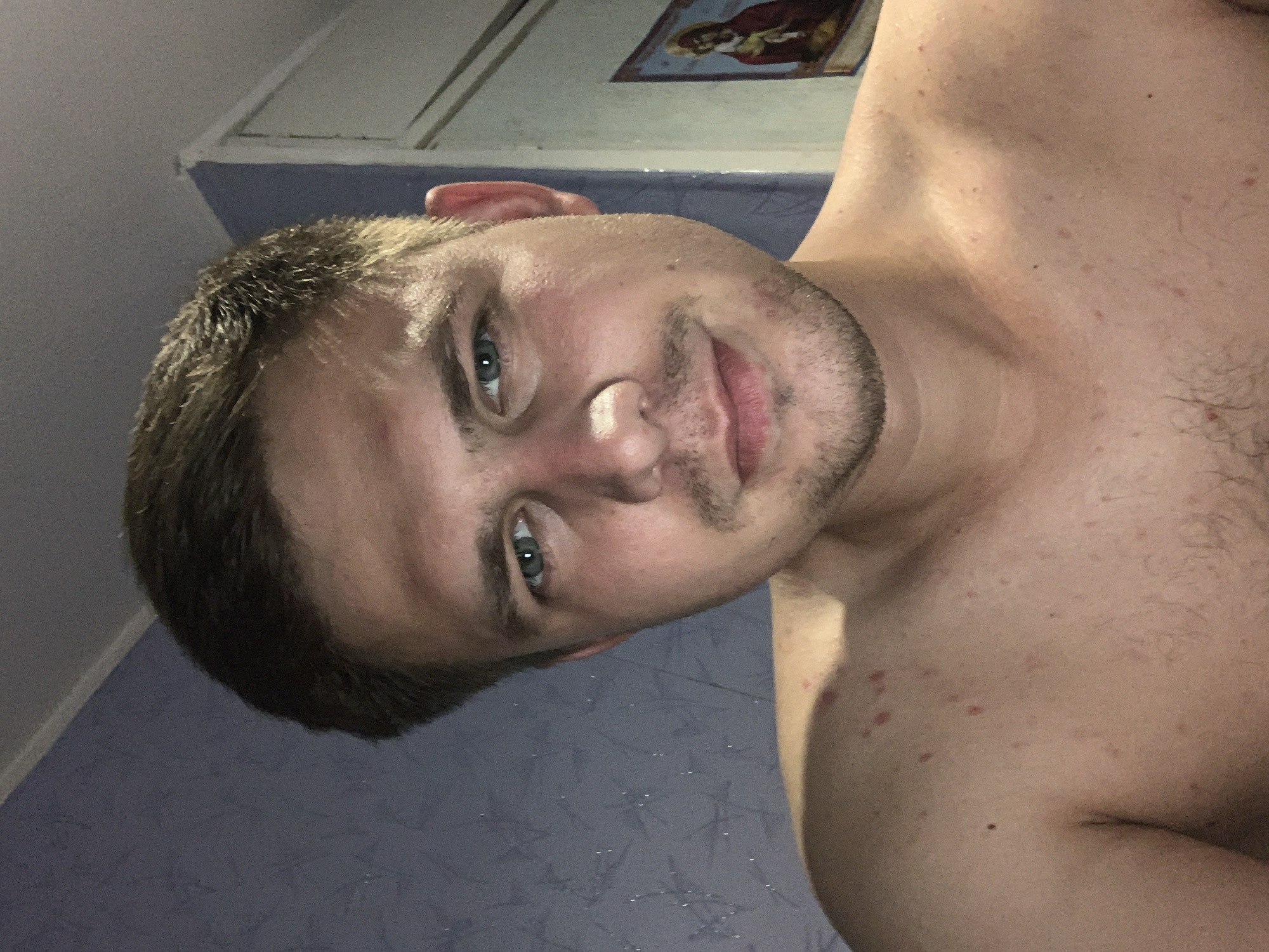 A close-up, shoulder-up selfie of a light-skinned, adult male with short, brown hair and light blue eyes. He is looking directly at the camera with a slight smile. He is shirtless, revealing his chest and shoulders, which have several red blemishes. He has a light stubble beard and mustache. In the background, a purple, textured wall is visible to the left, and a white door frame with a religious icon is visible to the right. The overall lighting suggests the photograph was taken indoors, possibly at night. The location is Unknown, Unknown.