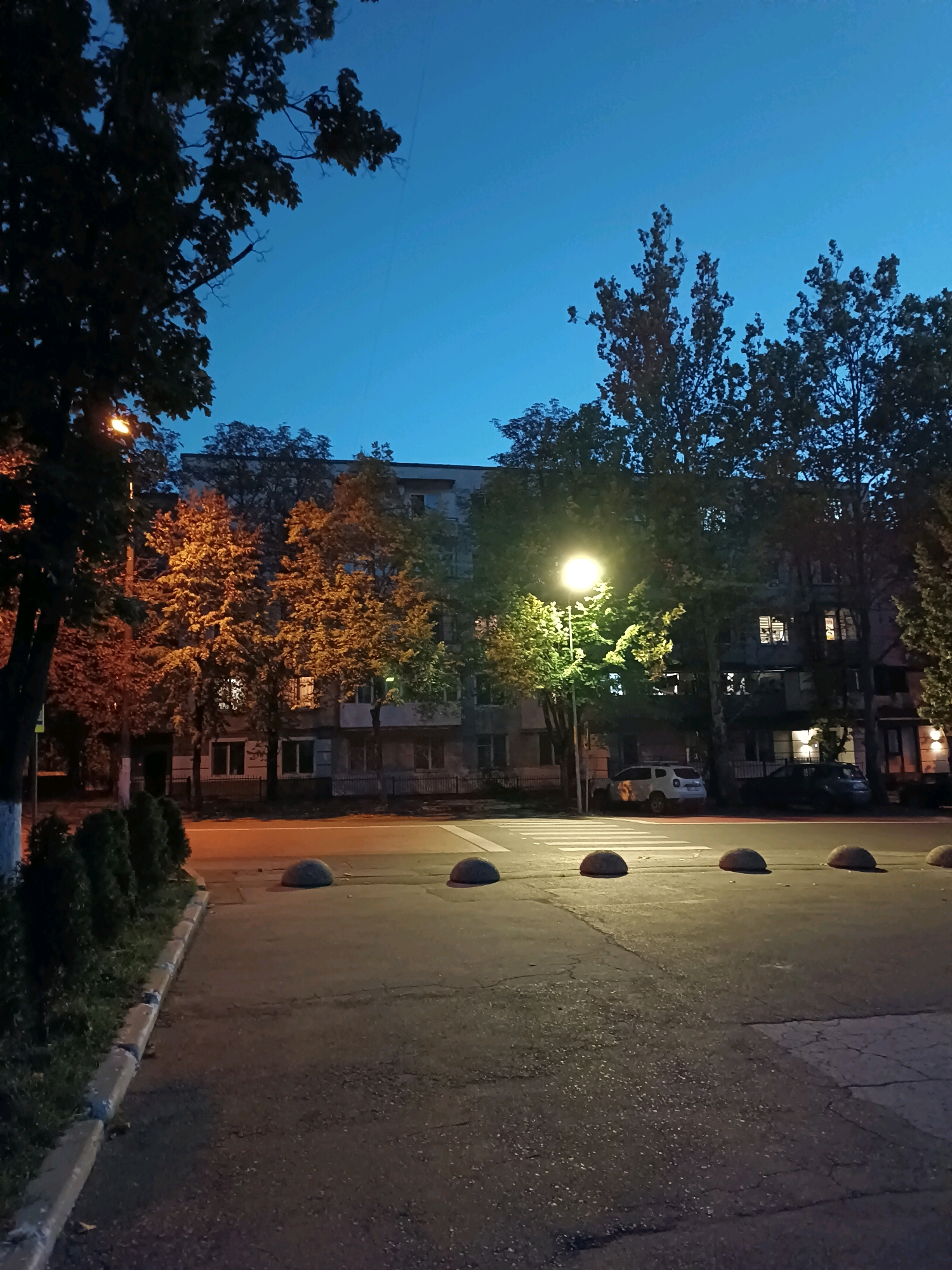 A street view in Chișinău, Moldova, at dusk. A light pole illuminates a pedestrian crosswalk, casting a warm glow on the surrounding trees and apartment buildings. The sky is a deep twilight blue, with the faintest hints of clouds. Cars are parked along the street, and concrete bollards line the crosswalk. The scene suggests a quiet evening in a residential area.