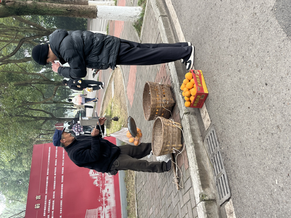 The image depicts two men outdoors. One man, wearing a black jacket and beanie, appears to be bringing something to his mouth. Nearby, a vendor is selling oranges from baskets and a cardboard box labeled with Chinese characters, using a makeshift scale. A sign with red text and an image of a building is in the background. People are strolling along a path lined with trees. The text on the sign includes the phrases "中国", "新中国", "发展史", "社会主义", "胜利", and "100年".