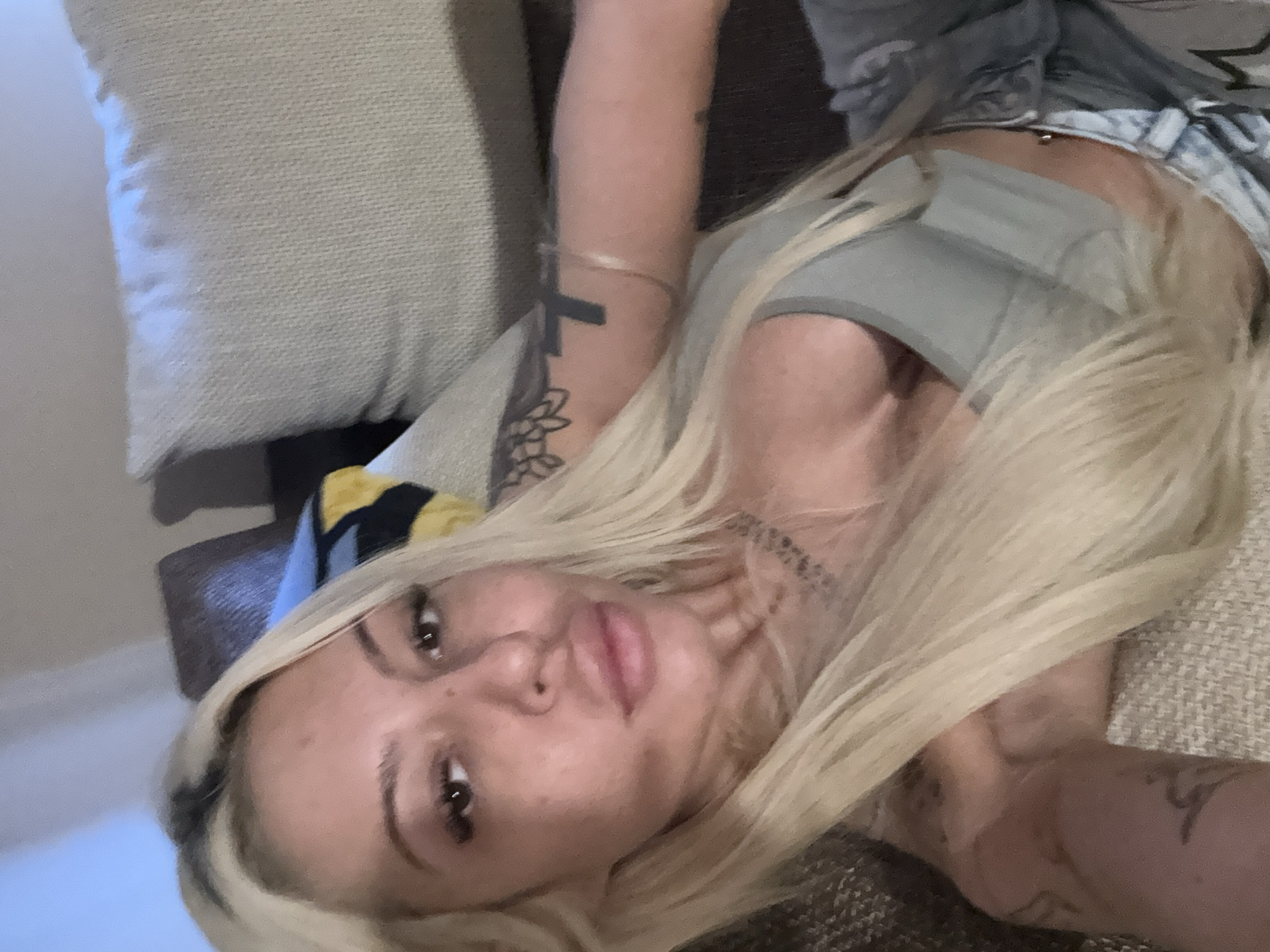 The image shows a young woman with long, straight blonde hair, featuring darker roots, lying down and looking directly at the camera. Her face is prominent, characterized by full lips, dark eyebrows, and dark eyes, with visible freckles across her nose and cheeks. She wears a light, possibly gray, crop top or bra, revealing a navel piercing. Another patterned garment is visible around her waist, suggesting shorts or a tied shirt.
Multiple tattoos are visible on her body. On her right arm, a black cross tattoo and a floral or mandala-like design are present. A thin metallic bracelet adorns her wrist. A geometric tattoo can be seen on her left arm. A script or patterned tattoo is also visible on her neck/collarbone area, but its text is not legible.
The setting appears to be indoors, likely a home, with soft, neutral-toned furnishings, including a textured pillow and a sofa or bed. The lighting is soft and even, consistent with a daytime indoor environment. The woman's expression is neutral and direct. The photograph is taken from a slightly overhead, tilted angle, resembling a selfie. There is no discernible legible text within the image apart from the non-decipherable neck tattoo. No visual cues within the image confirm the provided location context of Plovdiv, Bulgaria.