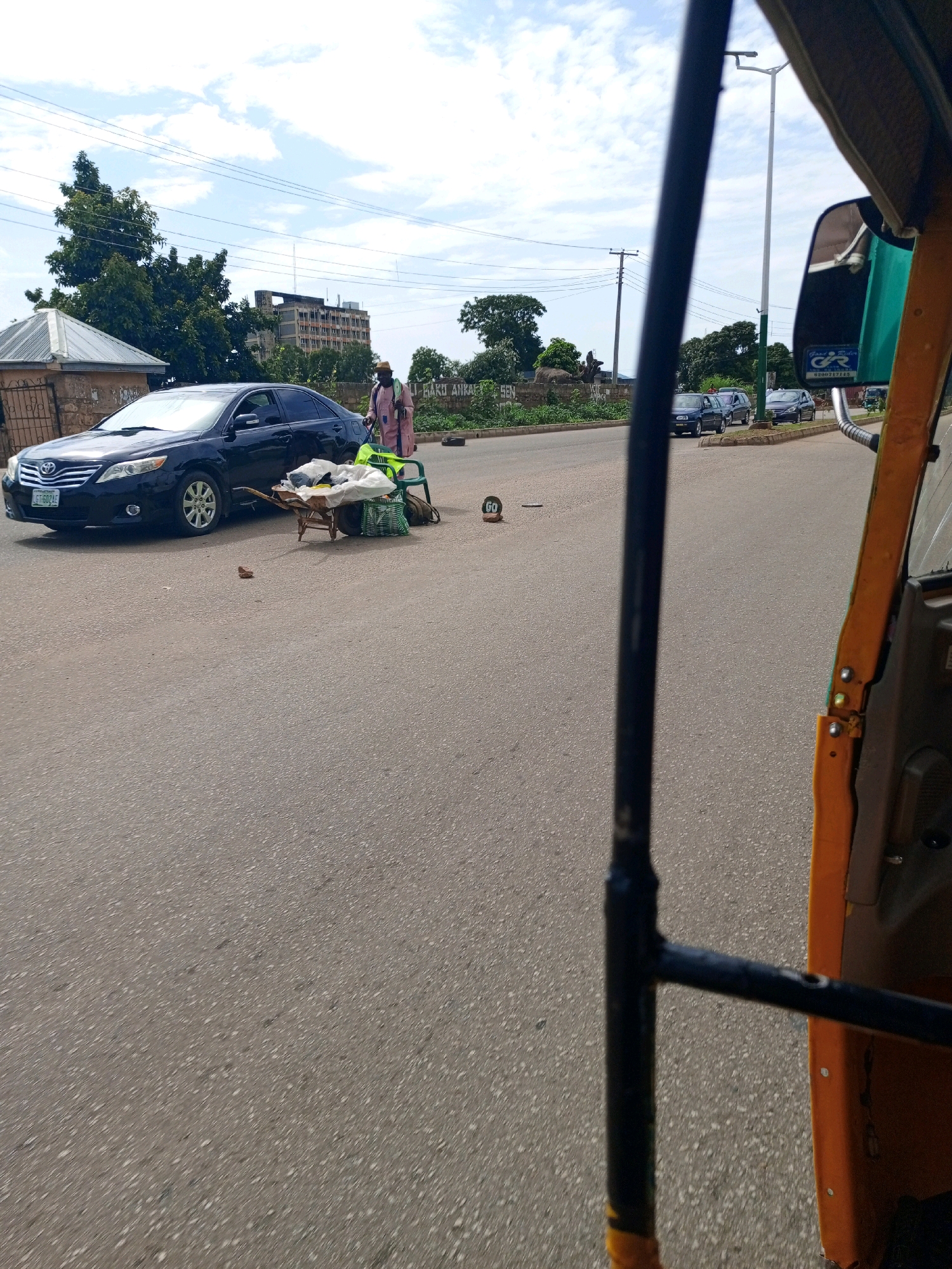A medium-angle outdoor shot captures a street scene in Jos, Nigeria, under a partly cloudy sky. In the foreground, a portion of an orange and yellow auto-rickshaw is visible on the right side of the frame, obscuring the full view of the street.
The primary subject is a person dressed in a pink outfit and a straw hat, standing next to a wheelbarrow. The wheelbarrow contains several items, including white plastic bags and a green plastic chair. The person appears to be attending to the wheelbarrow.
To the left of the person and wheelbarrow, a dark-colored sedan is parked. The car has a license plate with the inscription "GT 692 AE". Behind the car, a building with a metal roof structure is visible. Further in the background, there are trees, overhead wires, and a multi-story building.
On the road, a circular sign with the word "GO" is placed on its side. The road surface is asphalt with visible white aggregate. The overall impression is that of a typical street scene in a Nigerian city, with elements of commerce and daily life.