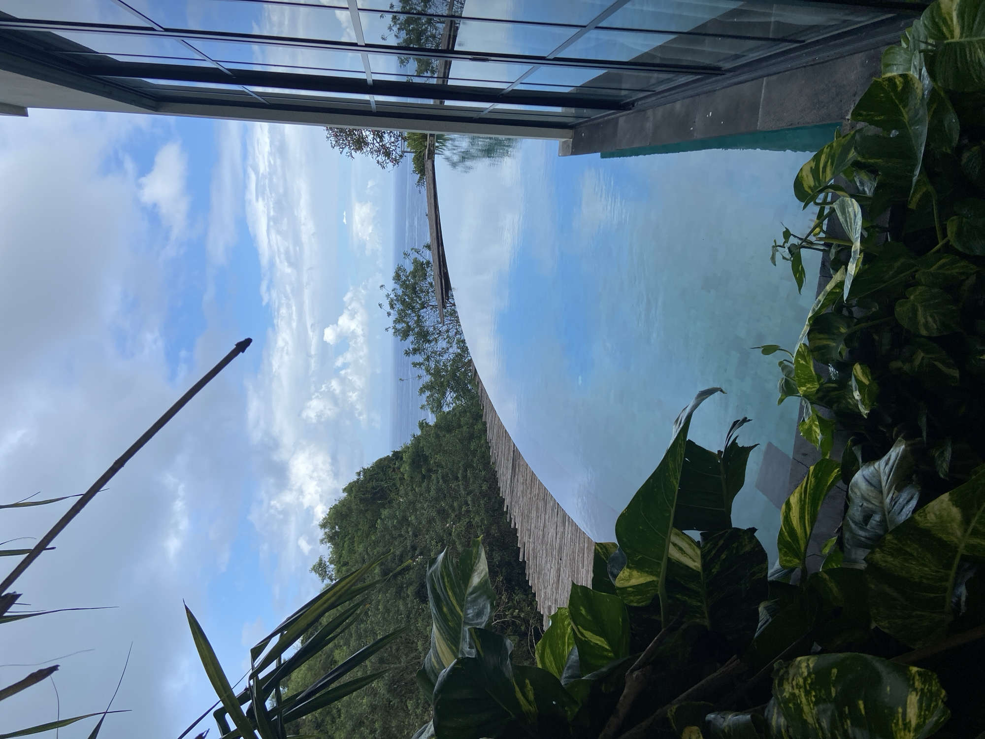 The image depicts a view from what appears to be a building in Kuta Selatan, Indonesia, overlooking a swimming pool with a seemingly curved edge that visually blends into the horizon. The sky is visible with scattered clouds, reflected in the pool's surface. Lush green vegetation is present in the foreground, suggesting a tropical environment. The building has a contemporary design with large glass windows. The overall scene suggests a high-end resort or private residence with an infinity pool feature.