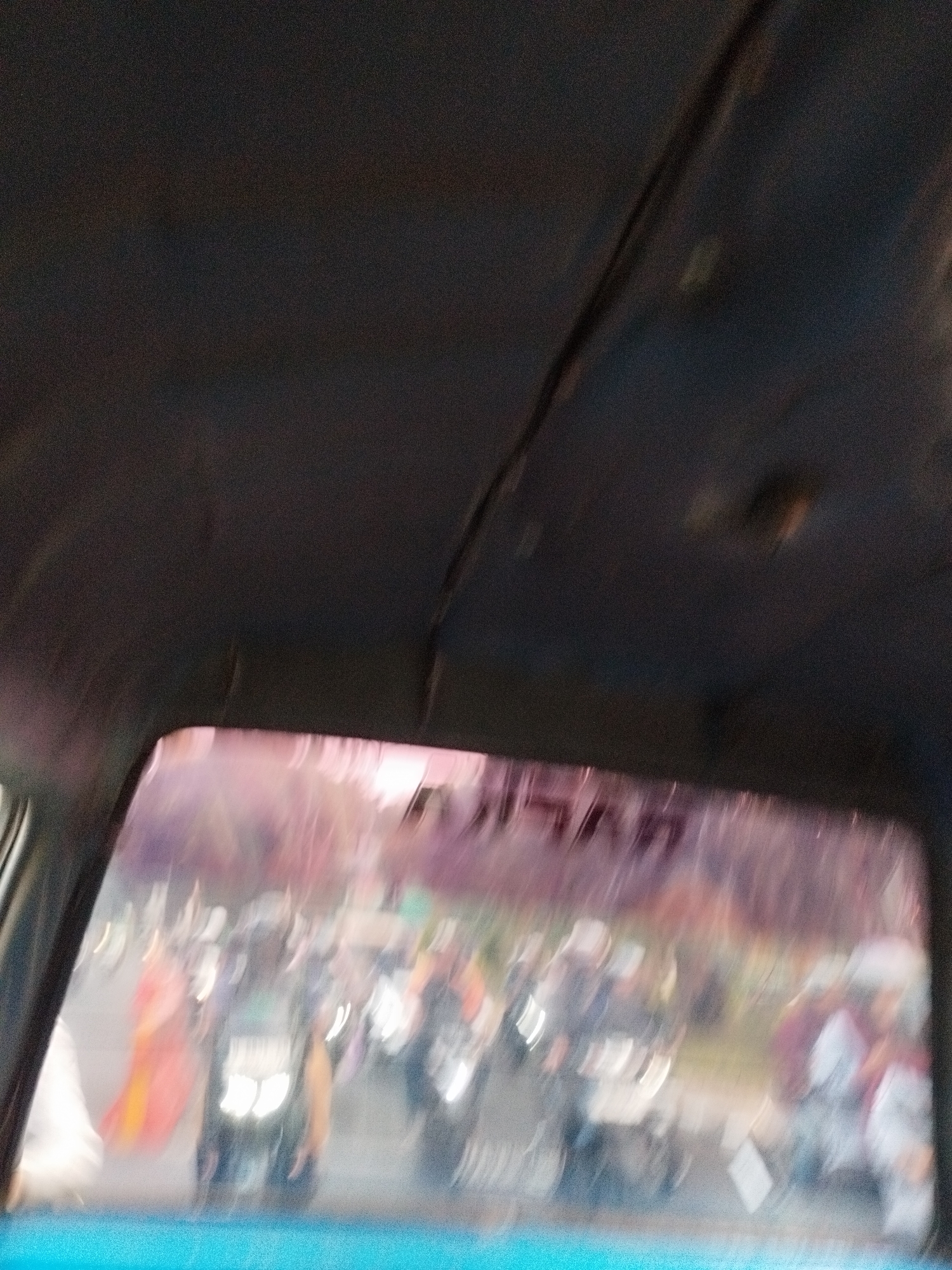 The image shows a view from inside a vehicle looking out through the windshield. The windshield is tinted with a pinkish hue, and there is text visible on the top of the windshield, though it is blurred and difficult to read. Outside the vehicle, the scene appears to be a street with several motorcycles and people. The lighting suggests it is either dawn or dusk. The background is blurred, but it looks like there are trees and buildings. The image is grainy and out of focus, making it difficult to discern fine details. It is possible the image was taken in Surabaya, Indonesia, given the presence of motorcycles and the general street scene.