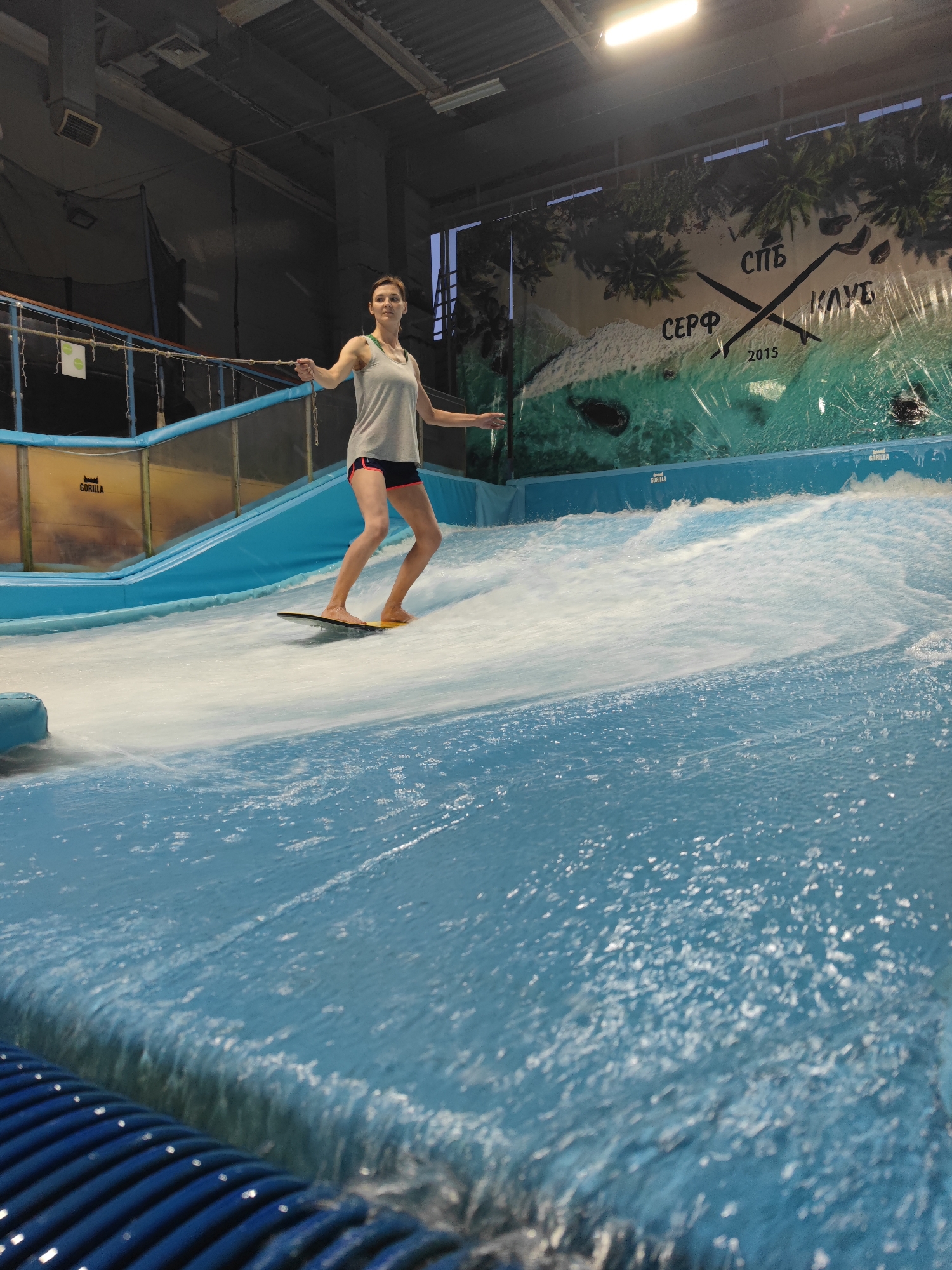 A young woman is depicted surfing on a stationary wave machine. She is standing on a yellow board with a black edge, wearing a grey tank top and dark shorts with pink trim. Her stance is balanced, suggesting she is actively engaged in surfing. The setting appears to be an indoor surf park, evident from the artificial wave, the enclosed space, and the lighting.
The background features a large mural depicting a tropical beach scene with palm trees, sand, and turquoise water. The mural also has text on it, including "СПБ КЛУБ" which likely refers to "Saint Petersburg Club" and "СЕРФ 2015" meaning "Surf 2015". This suggests the location is a surf club in Saint Petersburg, established in 2015.
The water is a vibrant blue and is actively moving, creating the illusion of waves. The lighting is artificial, typical of an indoor environment, and appears bright. There are no indications of the time of day or weather conditions as it is an indoor setting. The woman's expression is neutral to slightly focused, showing concentration on her surfing activity. The overall mood is energetic and recreational, highlighting the enjoyment of water sports in an unconventional setting. The blue ribbed structure in the foreground is part of the water management system of the surf machine. Visible text on the yellow wall behind the woman states "GORILLA".
