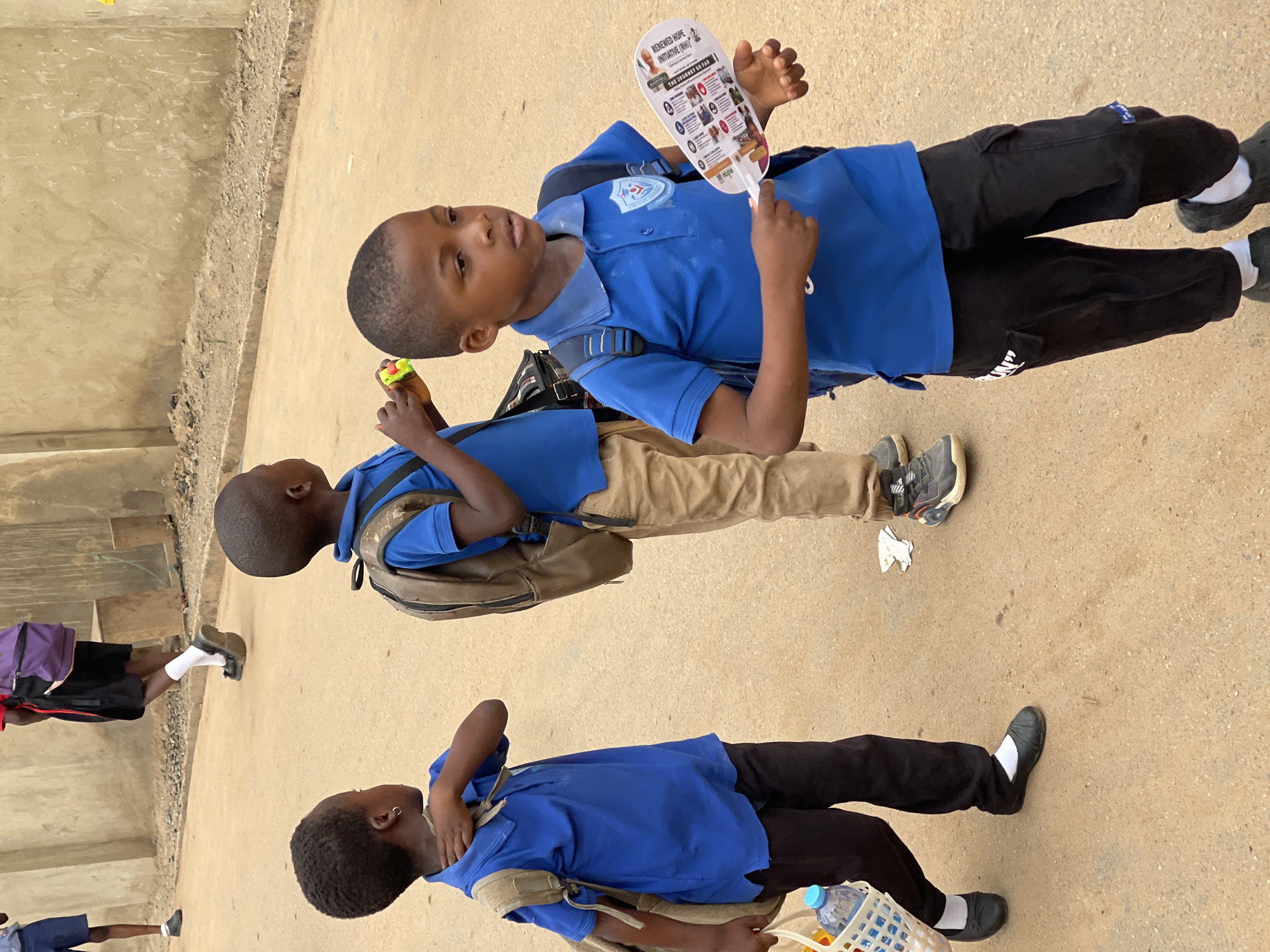 The image shows three black boys outdoors on a concrete surface, likely a street, in what could be Karmajiji, Nigeria. They are wearing blue shirts, presumably school uniforms, and backpacks. The boy at the top is holding a small fan with images and text on it. The boy in the middle is holding a small, colorful object. The bottom boy has his hand on his neck and appears to be carrying a white basket with a water bottle inside.