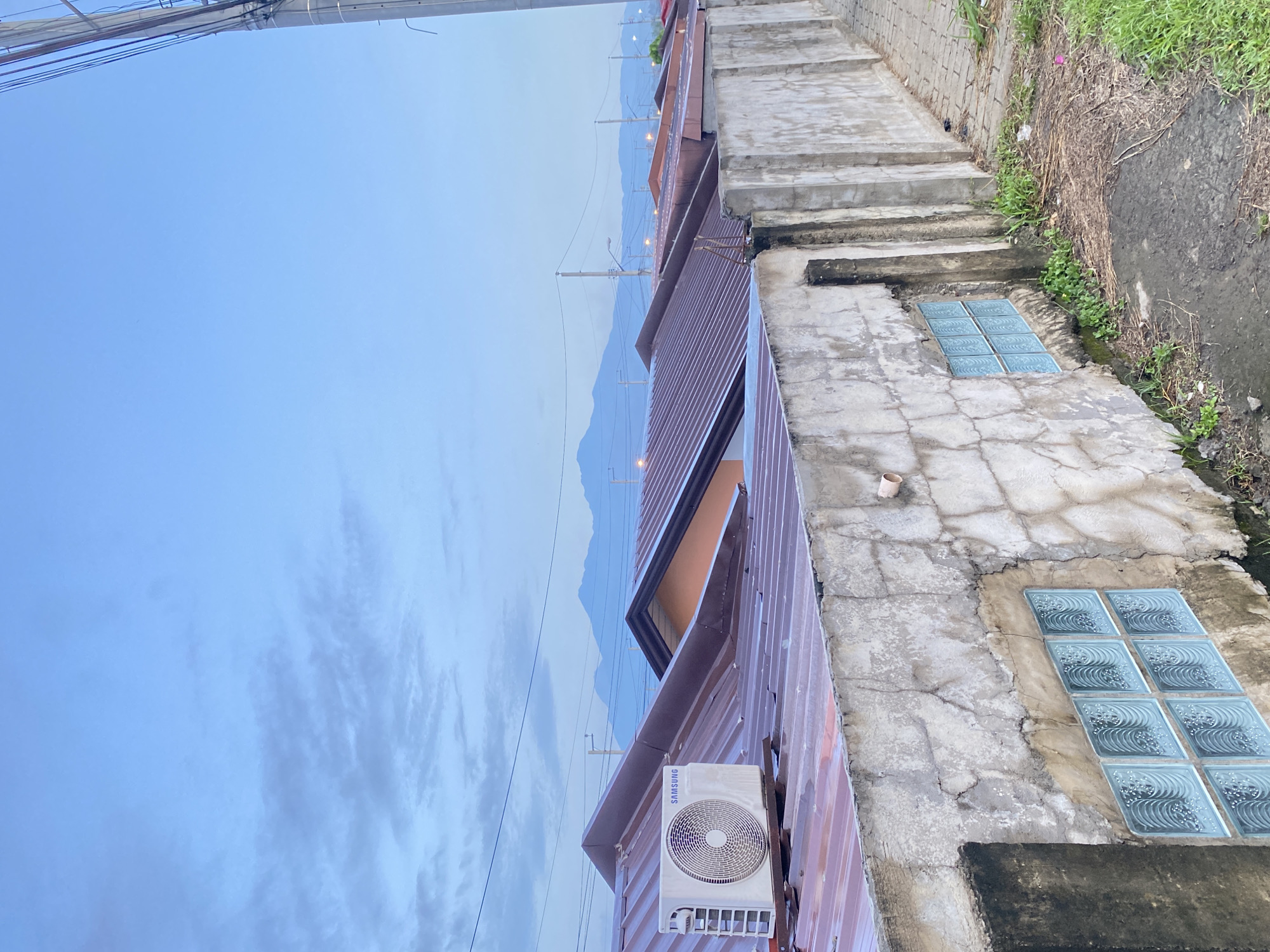 The image, correctly oriented horizontally, captures a view over a residential area with mountains in the distance, likely during dawn or dusk. The perspective is slightly elevated, looking down across rooftops towards the horizon.
In the immediate foreground on the right, there's a textured concrete pathway or staircase, showing signs of wear and cracks. Adjacent to this path, two rectangular concrete structures are visible, each embedding several frosted glass blocks with a wave pattern. Beyond these structures, a patch of green grass and weeds grows along a rough concrete wall.
To the left, the corrugated metal roofs of several buildings dominate the midground. One building closer to the viewer features a white Samsung air conditioner unit mounted on its side. Further down the row of buildings, more rooftops are visible, and several utility poles with numerous electrical wires crisscross the frame, receding into the distance. Some small, distant lights are faintly visible among the buildings, suggesting artificial illumination.
The background features silhouettes of mountains, appearing hazy and soft against the sky. The sky itself transitions from a light, clear blue at the top to a slightly cloudier, paler blue closer to the mountain peaks, with wispy cirrus clouds present in the upper left portion. No people or specific activities are visible, presenting a quiet, panoramic view of the urban-natural interface. The only visible text is "SAMSUNG" on the air conditioner unit.