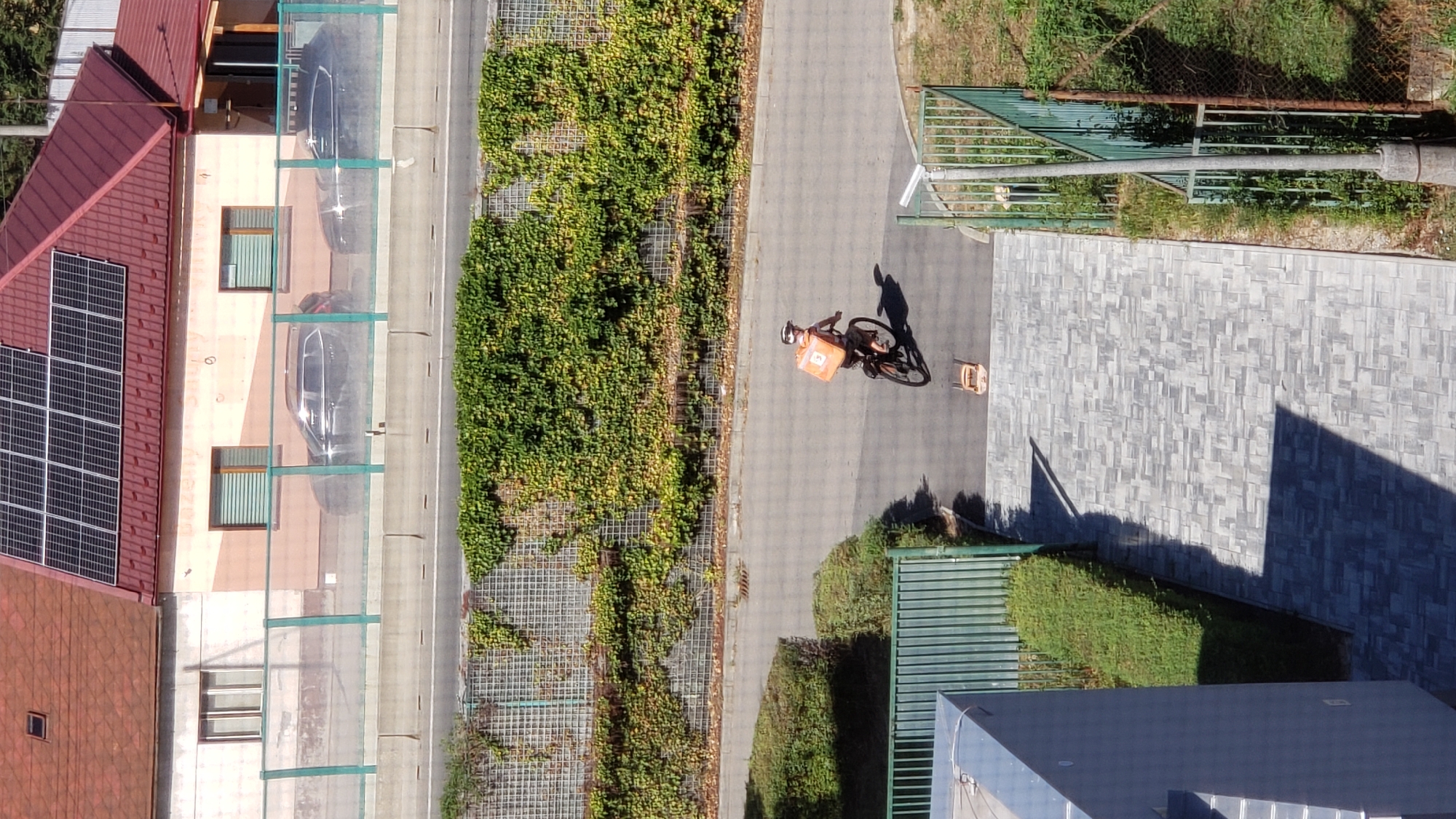 An aerial view captures a person on a bicycle, wearing a helmet and an orange delivery bag, riding on an asphalt road. The road is bordered by a grassy area and a fence. In the background, a building with a red roof and solar panels is visible, along with two parked cars. The scene appears to be in an urban or suburban setting, possibly Prešov, Slovakia, given the context. The lighting suggests it is daytime.