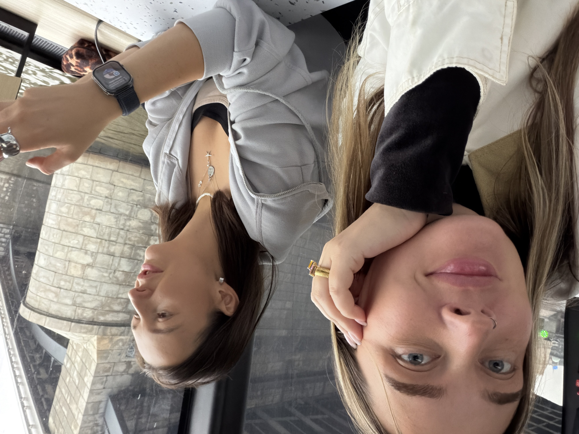 Two women are seated in an enclosed space, possibly a restaurant, with a view of the Moscow Tower. The woman on the left wears a light grey hooded sweatshirt, a necklace with a star charm, a silver ring, and a smartwatch. The woman on the right wears a jacket with a black undershirt and a large gold ring. The background shows the architecture of the Moscow Tower visible through a window.