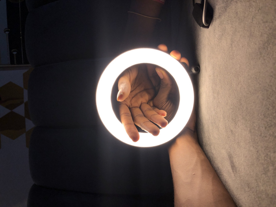 The image displays an interior scene, reportedly located in Tema, Ghana, focused on a brightly illuminated white LED ring light. A human hand with a medium-dark skin tone is positioned inside the glowing ring, palm partially open with fingers slightly splayed. This hand is being held or presented by an individual, whose wrist with a medium-dark skin tone and a light-colored bracelet is visible on the right side of the frame, partially obscured by the ring light. Another human forearm, with a lighter skin tone, extends from the left side into the lower-left foreground, resting on a textured light-grey surface. This same surface supports the ring light and the hands. The background is dimly lit and out of focus, revealing dark, possibly padded, shapes consistent with furniture like a sofa, and faint hexagonal patterns on a wall in the upper left quadrant. The ring light acts as the primary light source, brightly illuminating the central hand and the immediate foreground while casting the surrounding environment into shadow.