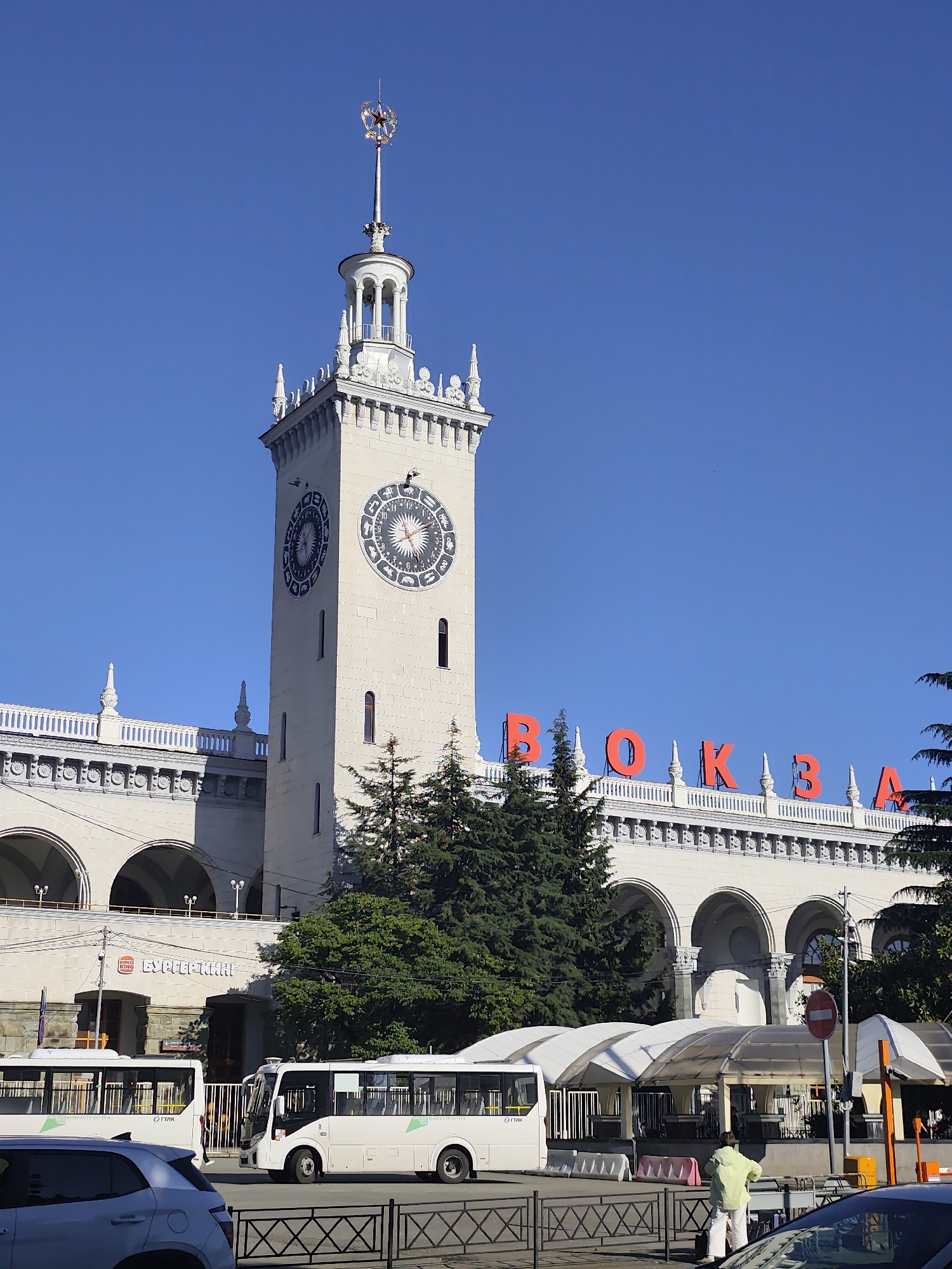 This image captures the grand train station in Sochi, Russia, on a clear, sunny day. The prominent, light-colored building, designed in the Stalinist Empire style, features a central clock tower topped with a spired cupola crowned by a golden star with red accents. Two large clocks on the tower display intricate, possibly zodiacal, symbols. Large red Cyrillic letters spelling "BOKZAЛ" (Vokzal, meaning "station") are affixed to the main facade of the building.
In the foreground, a street scene unfolds with various activities. Several white mini-buses are visible, two prominently in the mid-ground. One bus displays "ИНФРАСТРУКТУРА" (Infrastructure) and "ГТЛК" (GTLC) text, while another has "СИТИК" (Citic). A white car is partially visible on the left, and another on the right. Pedestrians include a person in a light green jacket and white pants standing near a black metal fence and white barriers on the right.
Arched entrances are visible at the base of the building, with a "БУРГЕР КИНГ" (Burger King) sign above one entrance. On the right side of the street, white tent-like structures with transparent roofs provide cover, possibly for an outdoor seating area or bus stop. The bright blue sky and strong sunlight suggest it is daytime, likely morning or early afternoon, with excellent weather conditions. The scene depicts a bustling transportation hub, combining historic architecture with modern commercial and transport elements.