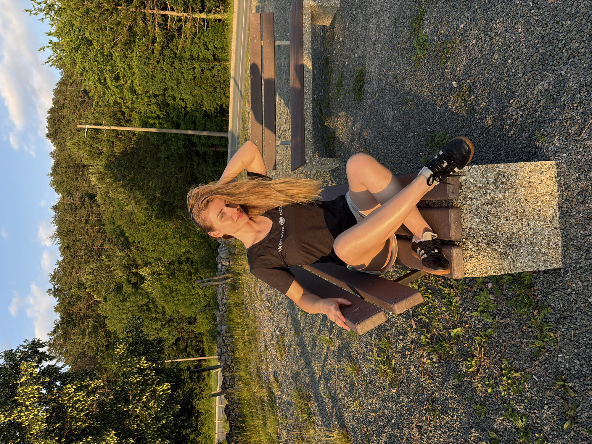 A full-body, eye-level, medium shot shows a young Caucasian woman with long blonde hair sitting on a park bench in a gravel-covered outdoor area. The woman is wearing a black t-shirt with white lettering and light-colored shorts. She is looking at the camera with her right hand on her head and her left hand resting on the bench. Her legs are crossed, and she is wearing black athletic shoes with white accents. The bench has a concrete base and wooden slats. In the background, there is a paved road, a wooden fence, a stone wall, and a dense forest. The sun is setting, casting long shadows and illuminating the scene with warm, golden light. The sky is blue with scattered white clouds.