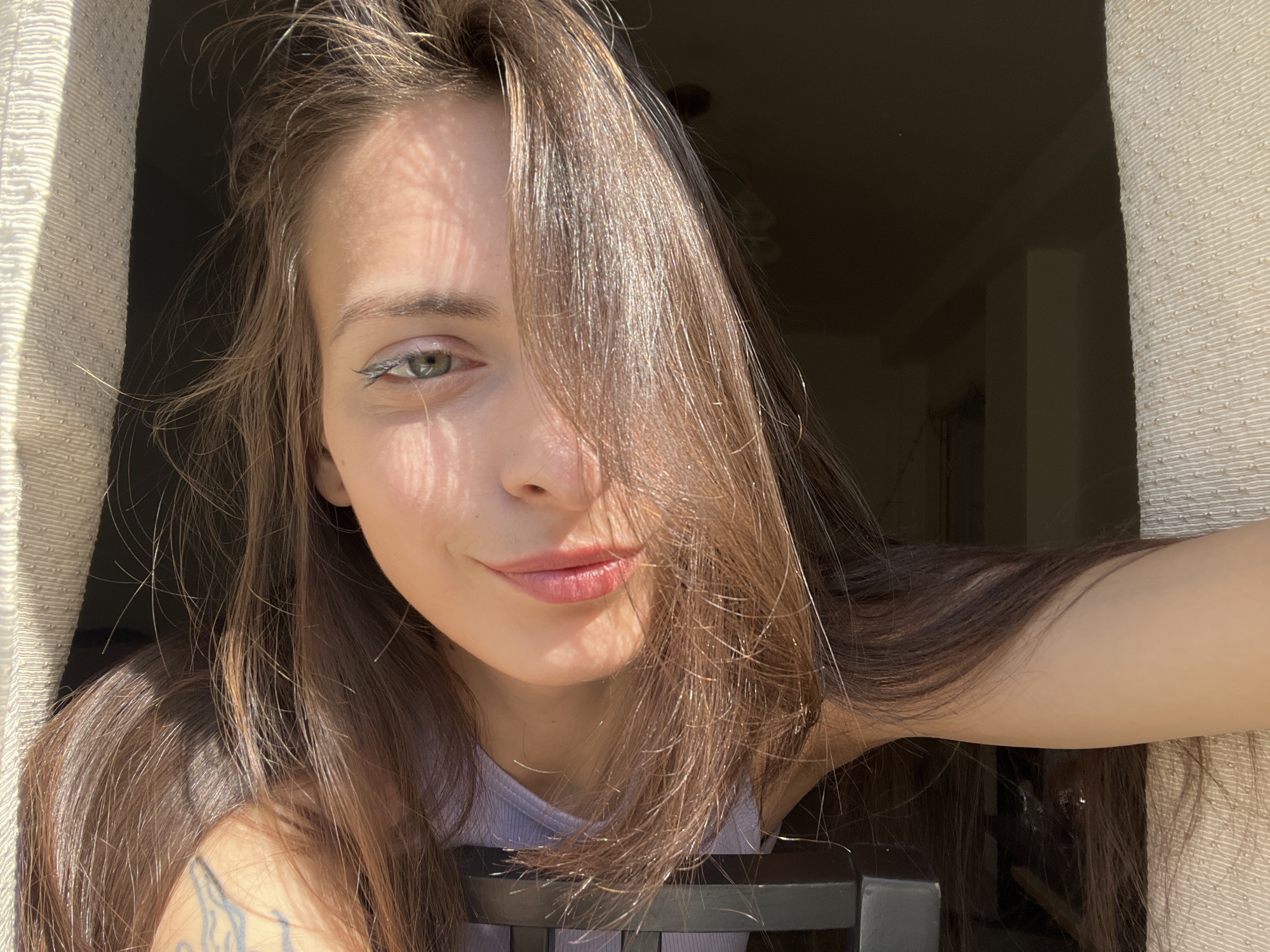 A young woman with long, dark brown hair and green eyes takes a close-up selfie indoors, bathed in bright sunlight, likely in Krasnoyarsk, Russia. Her hair is parted slightly off-center and frames her face, with strands catching the light. She has a subtle, natural smile on her face and is wearing light-colored eye makeup, possibly a white or light blue eyeliner. Sunlight filters in from the left, casting striped shadows across her face and hair, suggesting a window with blinds or textured curtains.
She is wearing a light-colored, possibly light purple or gray, ribbed tank top. A tattoo with a dark outline design is visible on her left arm, partially obscured by her hair. Her pose suggests she is leaning in towards the camera, possibly using her arm to hold the phone. To the left of the frame, a textured, light-colored curtain or blind is visible. The background to the right and behind her is dark and indistinct, indicating an interior space with less illumination. A dark, possibly black, chair back is visible in the lower central part of the image. The strong, direct light indicates a sunny day. There is no visible text in the image.