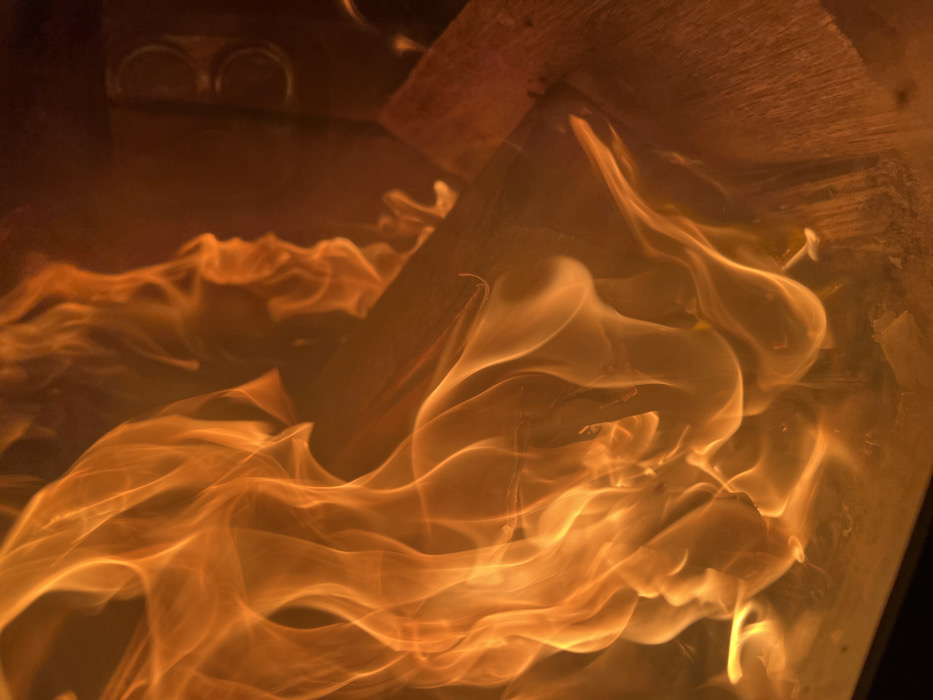 A close-up, vertical photograph depicts a contained fire in Sedlartsi, Bulgaria. The image is dominated by vibrant orange and yellow flames, exhibiting dynamic, upward-flowing, and swirling patterns, with brighter areas indicating higher heat. Partially combusted wood logs, showing charred surfaces, are visible underneath and through the flames, serving as fuel. Towards the right side of the frame, a dark, vertical surface, possibly metallic and part of a firebox or stove, is discernible. This surface features two circular, metallic components, stacked vertically, which may be air vents or control elements of the fire enclosure. No individuals are present in the scene.