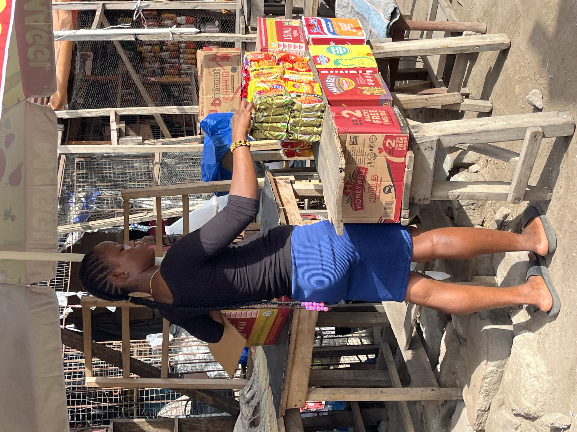 A street vendor is standing at a stall in Badagry, Nigeria. The vendor is a Black woman with long braids, wearing a black long-sleeved shirt and a blue skirt. She is reaching for a package of Indomie instant noodles on the stall. The stall is made of wood and displays a variety of instant noodle brands, including Indomie, Honeywell, and Twist Pasta. The street is crowded with wooden structures, and the ground is uneven.