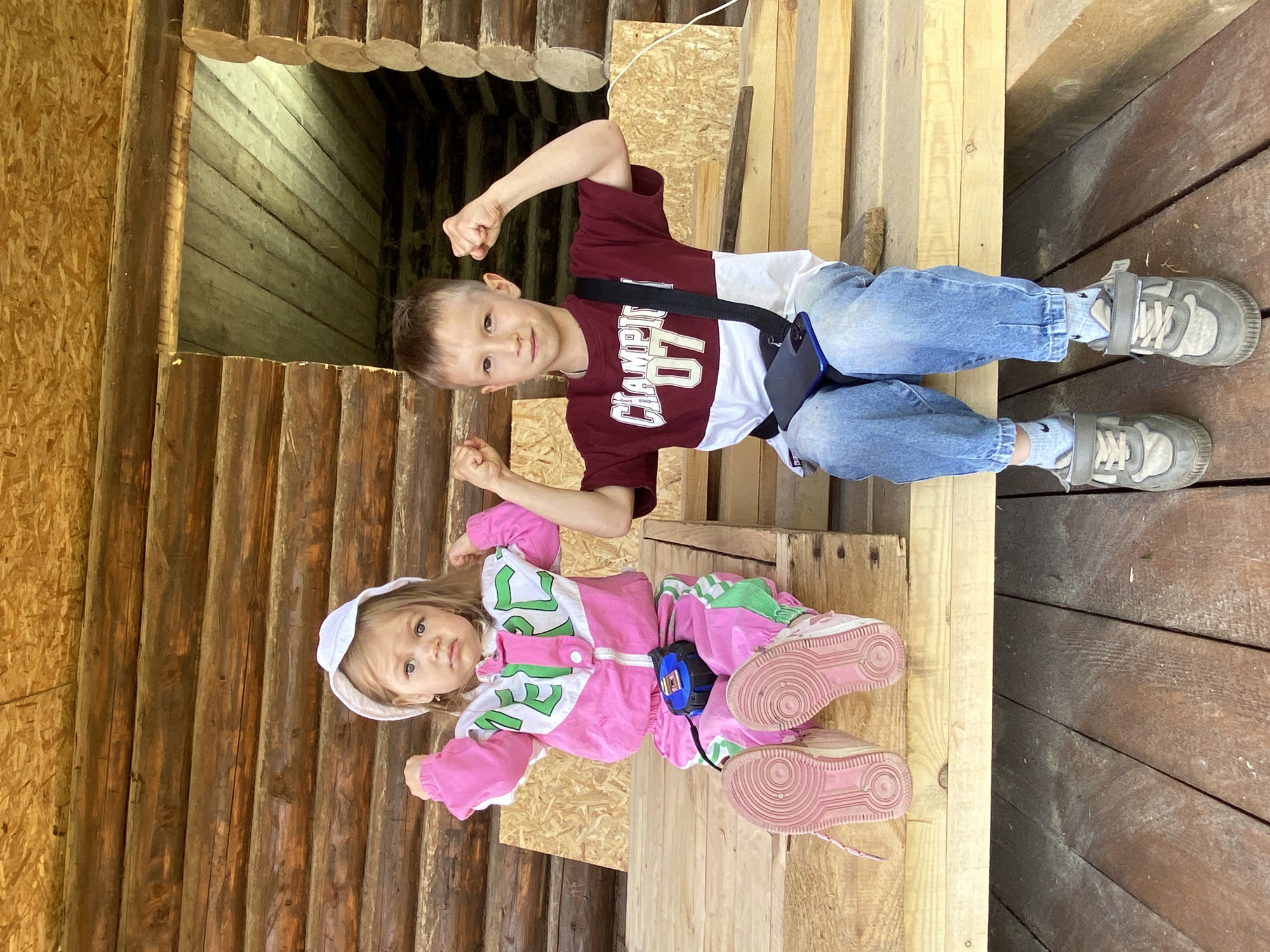 In the city of Chaikovsky, Russia, two children are seated on a raw wooden platform, posing with flexed biceps. The background features the unfinished log walls of a building and an overhead ceiling constructed from oriented strand board (OSB).
The child on the left is a young girl with light hair, wearing a white bucket hat and a pink and white tracksuit with green accents. The letters "MEPC" are visible on her top. A blue and black measuring tape is clipped to her tracksuit. She wears pink sneakers and has a serious expression, looking directly forward.
The child on the right is a boy with short brown hair, wearing a maroon, white, and red t-shirt displaying "CHAMPION" and "07." He wears light blue jeans, cuffed at the ankles, and grey and beige sneakers with Velcro straps. A black cross-body bag containing a dark blue smartphone is slung across his chest. He has a slight smile and looks directly at the camera.
Both children have their arms bent at the elbow, fists clenched, presenting a show of strength. Behind them, additional rough wooden planks and an open dark doorway leading into the log structure are visible. The scene is illuminated by natural light.