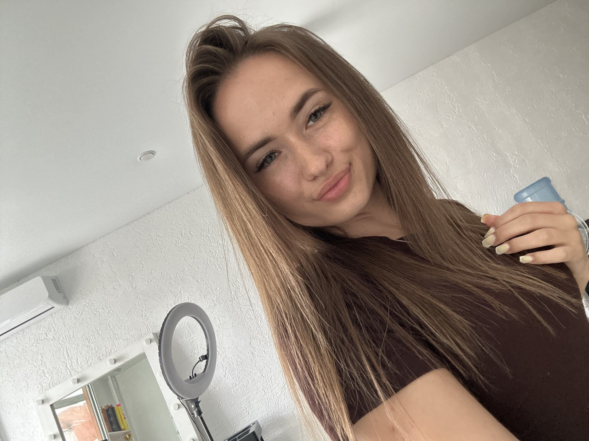 A young woman with long, light brown hair is taking a selfie in a room. She is wearing a dark brown top and has light-colored fingernails. She is holding a light blue object in her right hand. In the background, there is a ring light, a mirror with lights around it, and an air conditioning unit on a textured white wall. The ceiling is also white and has a circular light fixture.