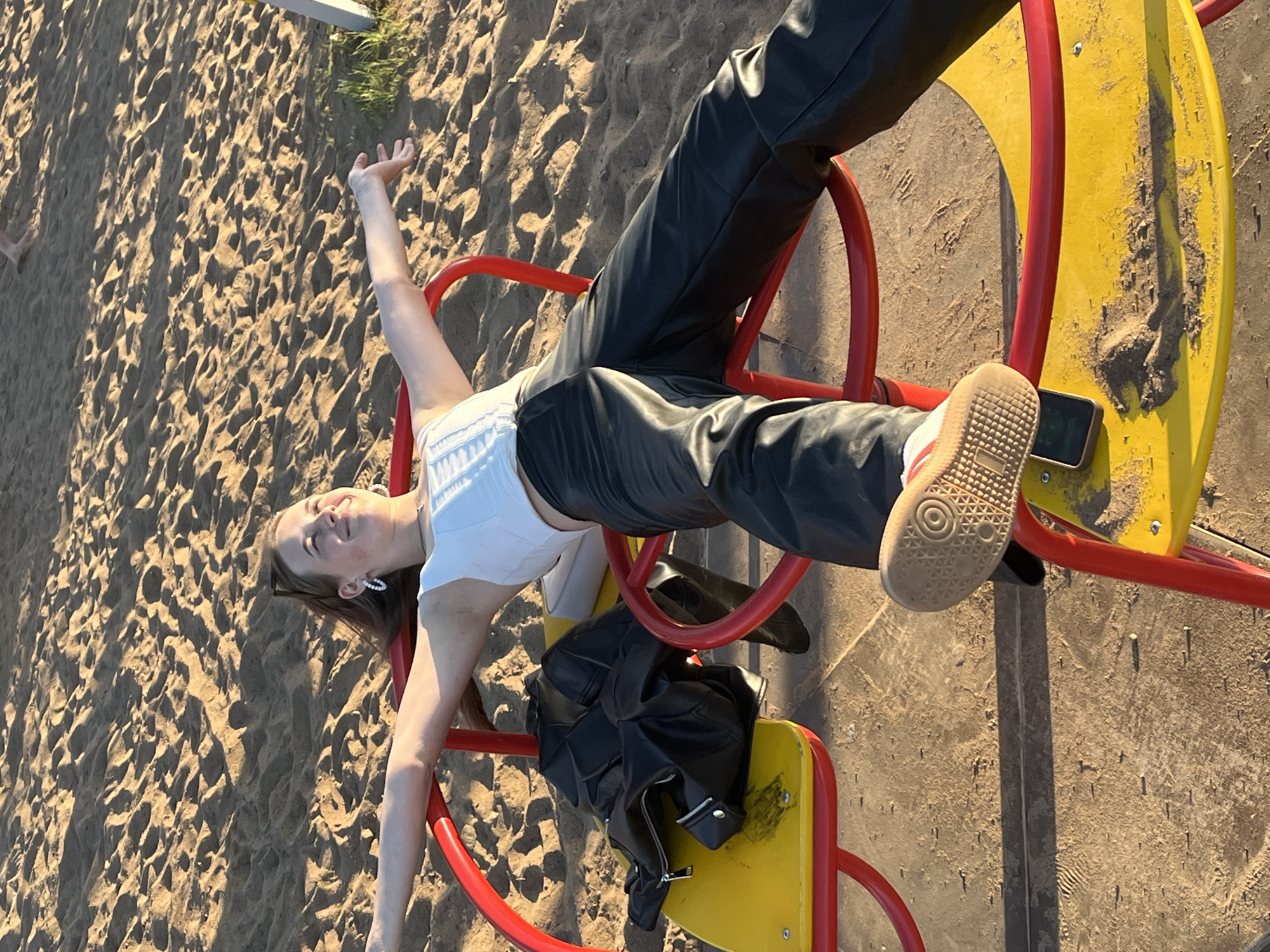 A young woman lies on a playground carousel located in a sandy area that could be near Фоки, Russia. She is wearing a white top and black leather pants. Her right arm is extended upwards, and her left arm is extended to the side. A black leather jacket and a cell phone are on the carousel seat near her feet. She appears relaxed. The carousel is painted yellow and red. The ground is covered in sand.