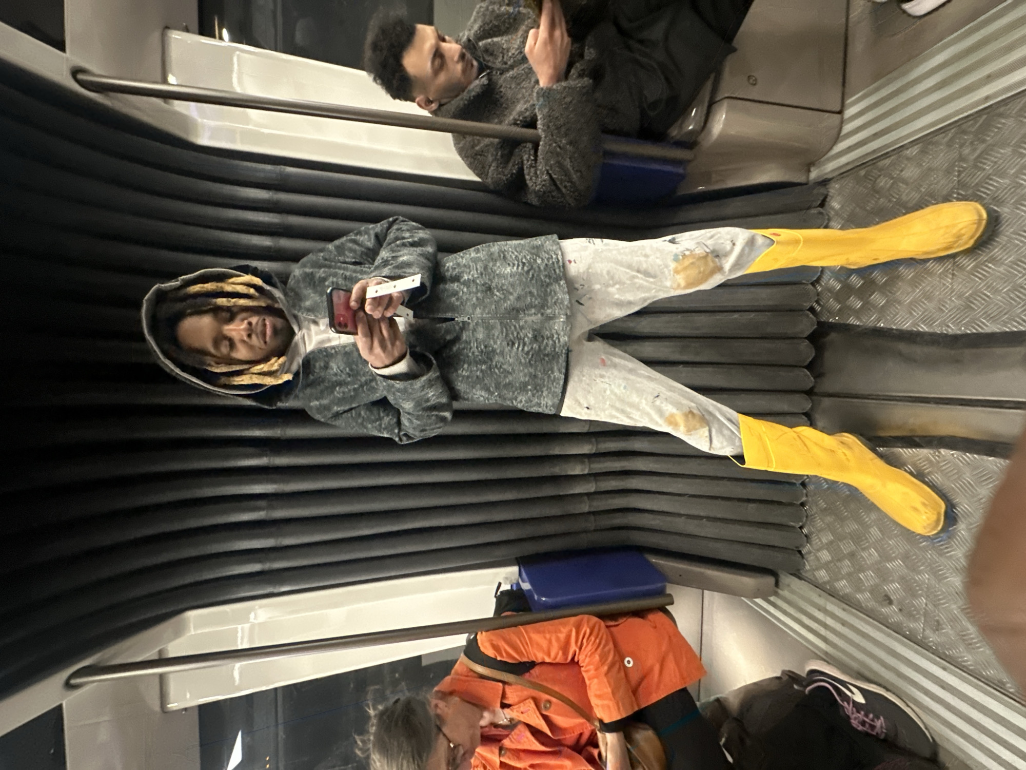 The image captures the interior of a public transportation vehicle, likely a metro or train in Paris, France, with several passengers. The scene is depicted as if the original image has been rotated 90 degrees counter-clockwise to an upright orientation.
In the foreground, a young man with dreadlocks wears a grey hooded jacket and light-colored pants that appear splattered with paint. He is holding a red smartphone with a white strap or lanyard attached, looking down at the screen. He wears distinctive, bright yellow rubber boots and has his legs stretched out, resting his feet against the ribbed side wall of the vehicle. His expression is calm.
Behind him, another young man is seated, dressed in a dark grey, textured jacket, also engrossed in his smartphone. Further down the aisle, partially visible, is a person wearing an orange jacket and glasses. A fourth person's leg and shoe are barely visible in the far right of the frame.
The interior of the vehicle features rows of seats, grey handrails, and windows reflecting ambient light. The floor has a metallic diamond plate pattern, and the lower walls are covered in dark, horizontally ribbed panels. A blue object, possibly a container or bag, is visible under one of the seats. The lighting is artificial, consistent with an indoor setting. Passengers are primarily engaged with their personal devices while traveling. No legible text is visible on any objects or screens.