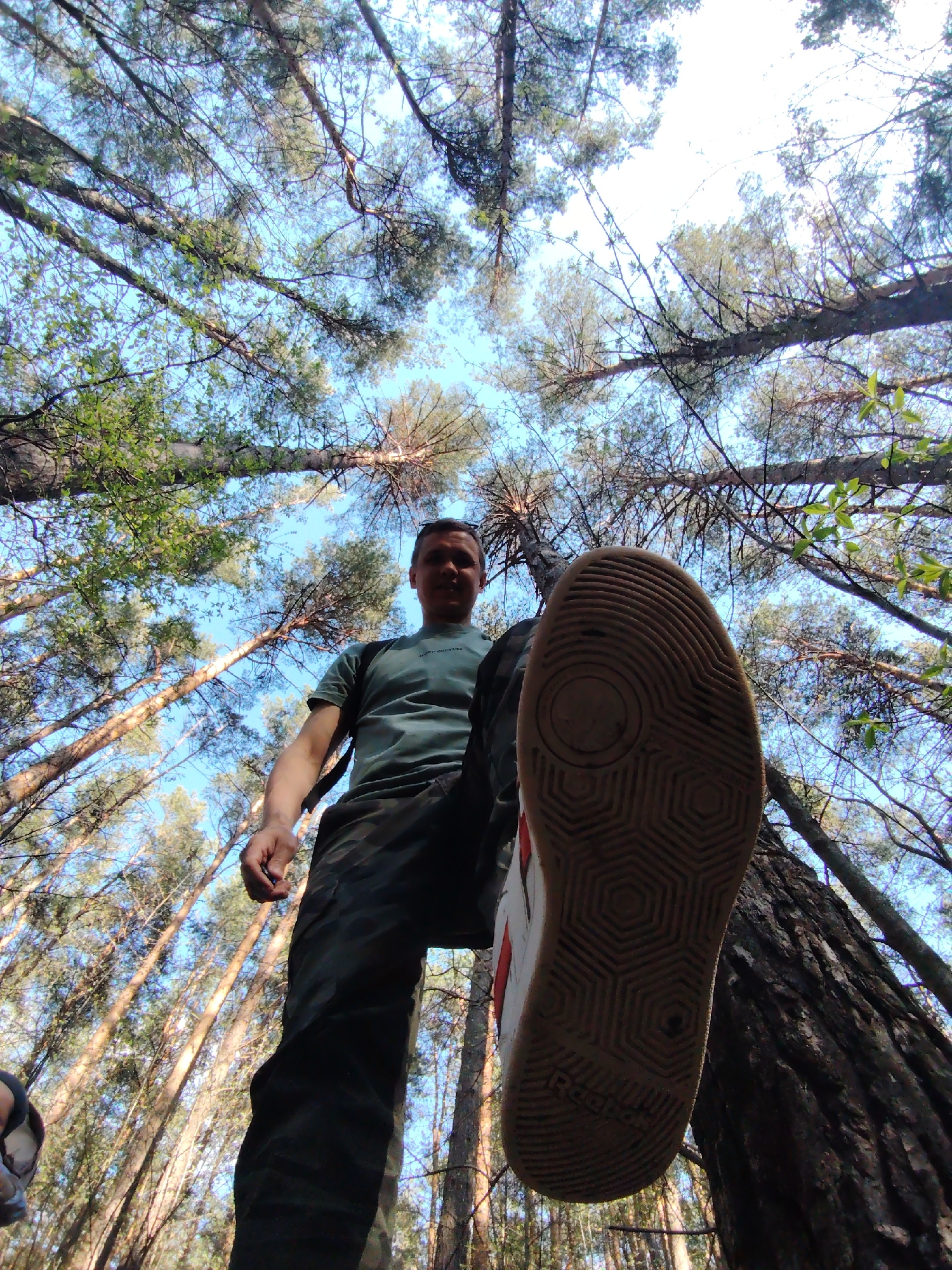 A low-angle shot captures a person in a forest setting, potentially in Chaikovsky, Russia. The person is wearing a green t-shirt, camouflage pants, and athletic shoes. The sole of one shoe is prominently displayed towards the camera, almost blocking the view of the person's face. Trees surround the individual, their branches reaching up toward a blue sky. The overall impression is one of being in a forest environment with someone standing overhead.