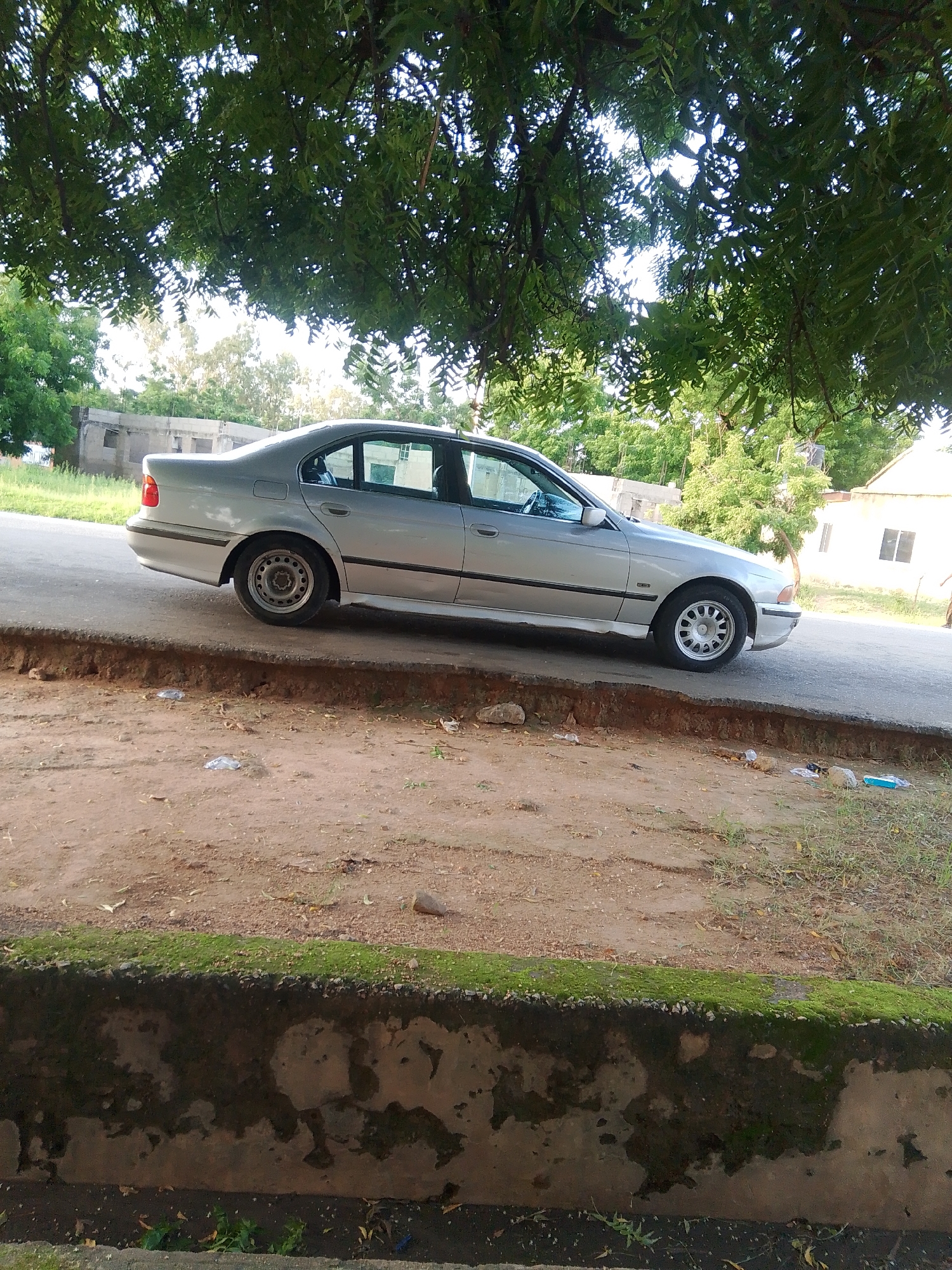 The image shows a silver sedan parked on an asphalt road. The car is a BMW, likely from the late 1990s or early 2000s, with a prominent side profile visible. It is parked under the shade of a large tree with dense green foliage, suggesting it is daytime. The road appears to be somewhat weathered, with a noticeable drop-off to a dirt and gravel area in the foreground.
In the background, several buildings are visible, some appearing partially constructed or in disrepair, while others are more complete. Green vegetation is abundant, with trees and grass dotting the landscape. The overall setting suggests an outdoor environment in a somewhat rural or developing area.
The lighting indicates it's likely daytime, possibly late afternoon given the angle of the sun creating dappled shade from the tree. There are no people clearly visible in the image, only the car and the surrounding environment. No text is discernible in the image. The scene is static, with the car parked and no apparent activity taking place. The mood is calm and ordinary.