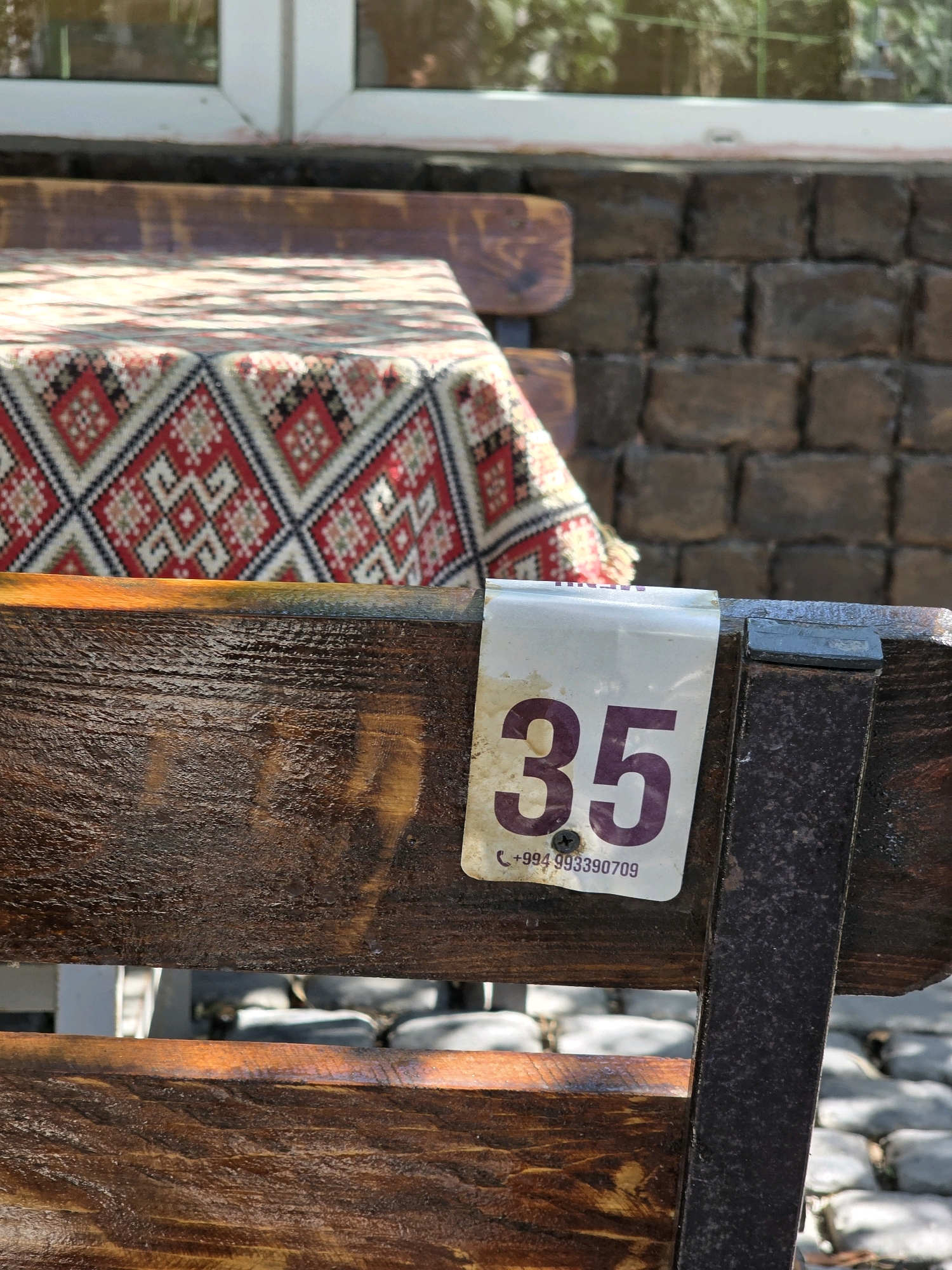 The image shows a close-up of a wooden bench with a table in the background covered with a patterned cloth. A white tag with the number "35" and a phone number "+994 993390709" is attached to the bench. The bench is weathered and appears to be outdoors, with a cobblestone ground visible. A brick wall and a window with a reflection are also visible in the background. The scene is likely an outdoor seating area of a restaurant or cafe in Qobustan, Azerbaijan.