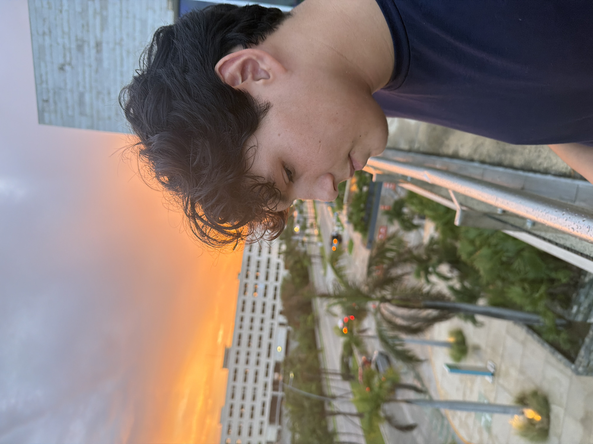 A young person with dark, wavy hair is seen from the side, looking down. They are wearing a dark blue t-shirt. The person is standing on a balcony or elevated walkway with a metal railing. In the background, a vibrant sunset paints the sky in shades of orange and yellow. Below the sunset, a large, white building, possibly a hotel or resort, is visible. Palm trees line a street with cars driving by. The environment appears to be urban, situated in Cancún, Mexico, characterized by tropical vegetation and modern architecture illuminated by the warm glow of the setting sun.