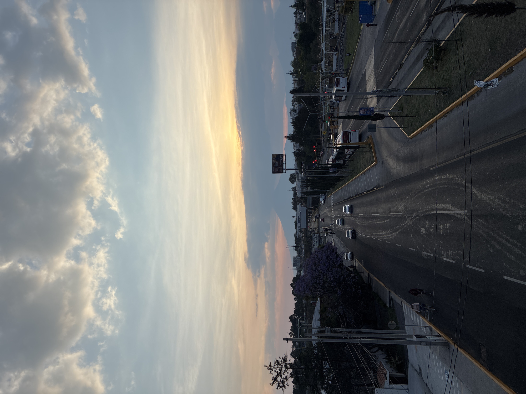 The image presents an elevated view of a road in San Bernardino Tlaxcalancingo, Mexico, at dusk. A multi-lane highway stretches into the distance, populated with cars and traversed by a cyclist. Traffic lights are visible along the roadside, with some displaying red. A billboard is present near an intersection. The sky exhibits colors ranging from pale blue to orange, indicating a sunset or sunrise. Power lines run along the foreground. Trees line parts of the roadside. The overall environment suggests a suburban or semi-urban setting.